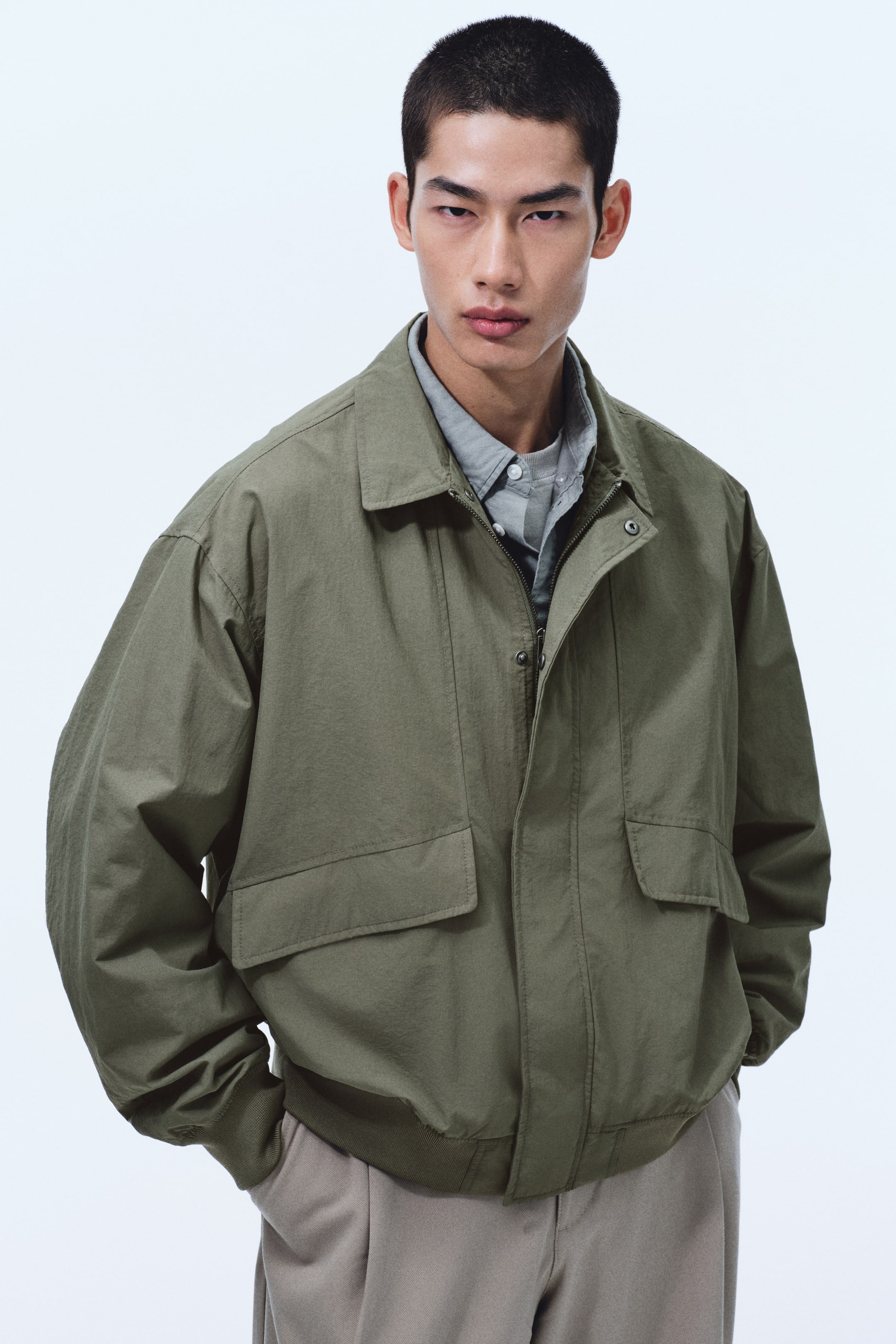 Xem ảnh lớn hơn: Chiếc áo bomber màu xanh ô liu nhìn từ phía trước có cổ nhọn và khóa kéo trước mở một phần, lộ ra chiếc áo sơ mi có cổ màu xám. Thiết kế có hai túi lớn dạng nắp ở phần trước dưới, cùng với bo gân ở cổ tay và lai áo.