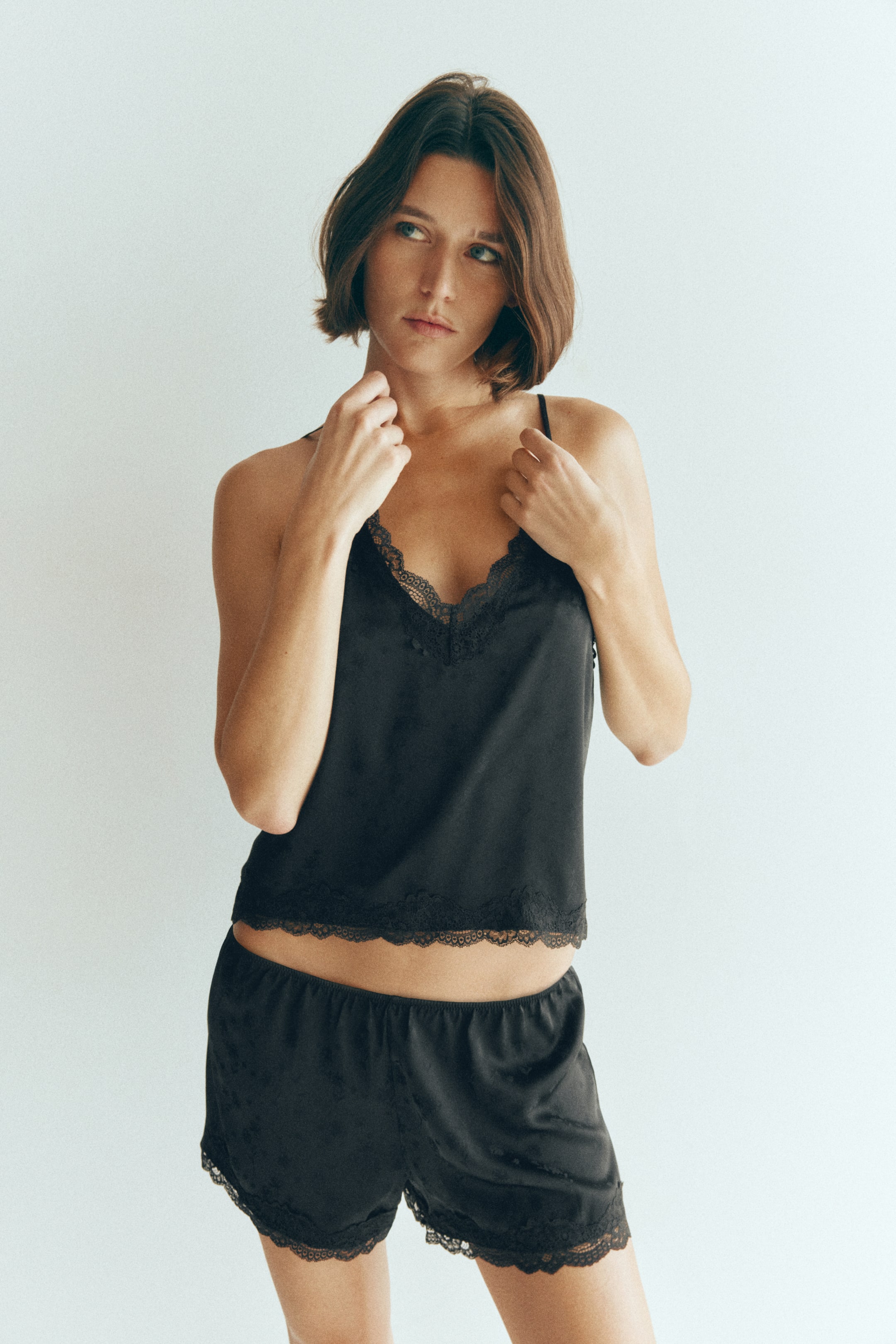 Visualizza immagine più grande: La modella indossa un pigiama nero in satin composto da una canotta con scollo a V e shorts coordinati con profili in pizzo. La modella tiene delicatamente tra le dita le sottili spalline a spaghetto del top vicino alla clavicola.