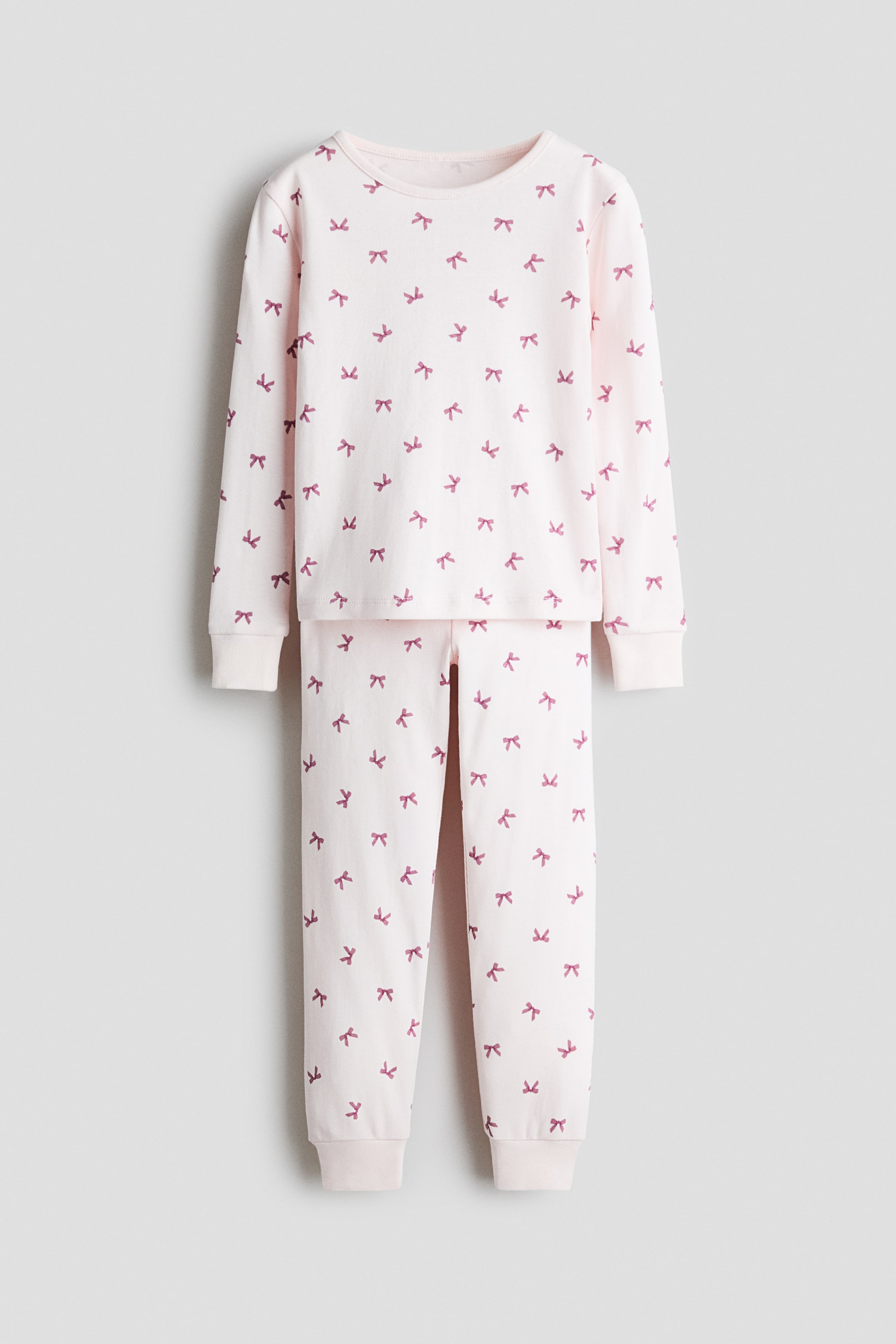 View larger image: A front-facing view of a light pink pajama set showing a matching long-sleeved top and pants with an all-over print of small, dark pink bows. The top features a round neckline and ribbed cuffs, while the matching pants taper to ribbed hems.