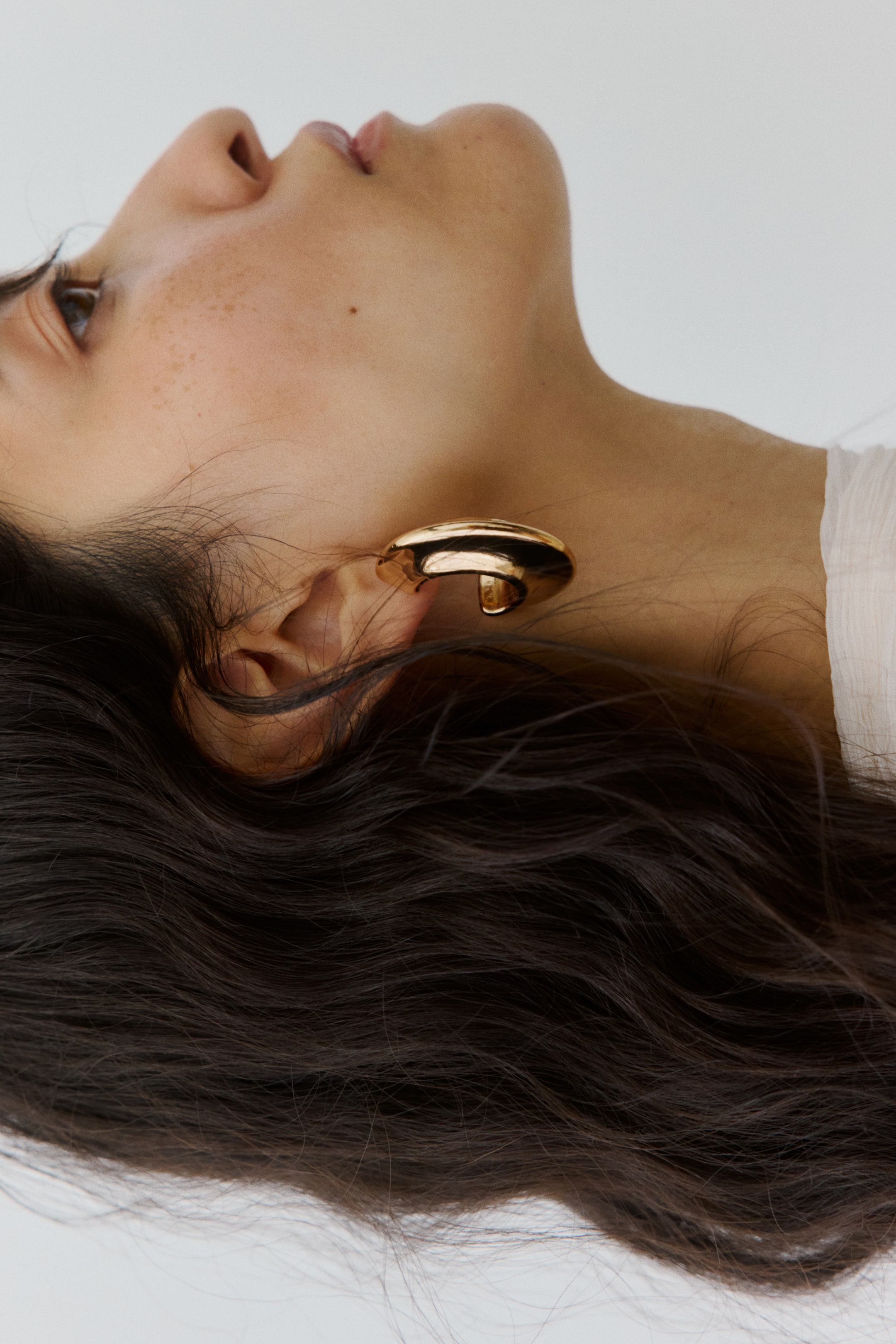 Agrandir l'image: Un manchette d'oreille dorée se courbe autour de l'hélix externe d'une oreille. Elle présente une forme sculpturale et polie avec une section supérieure plus large et arrondie qui s'effile en une bande plus fine.