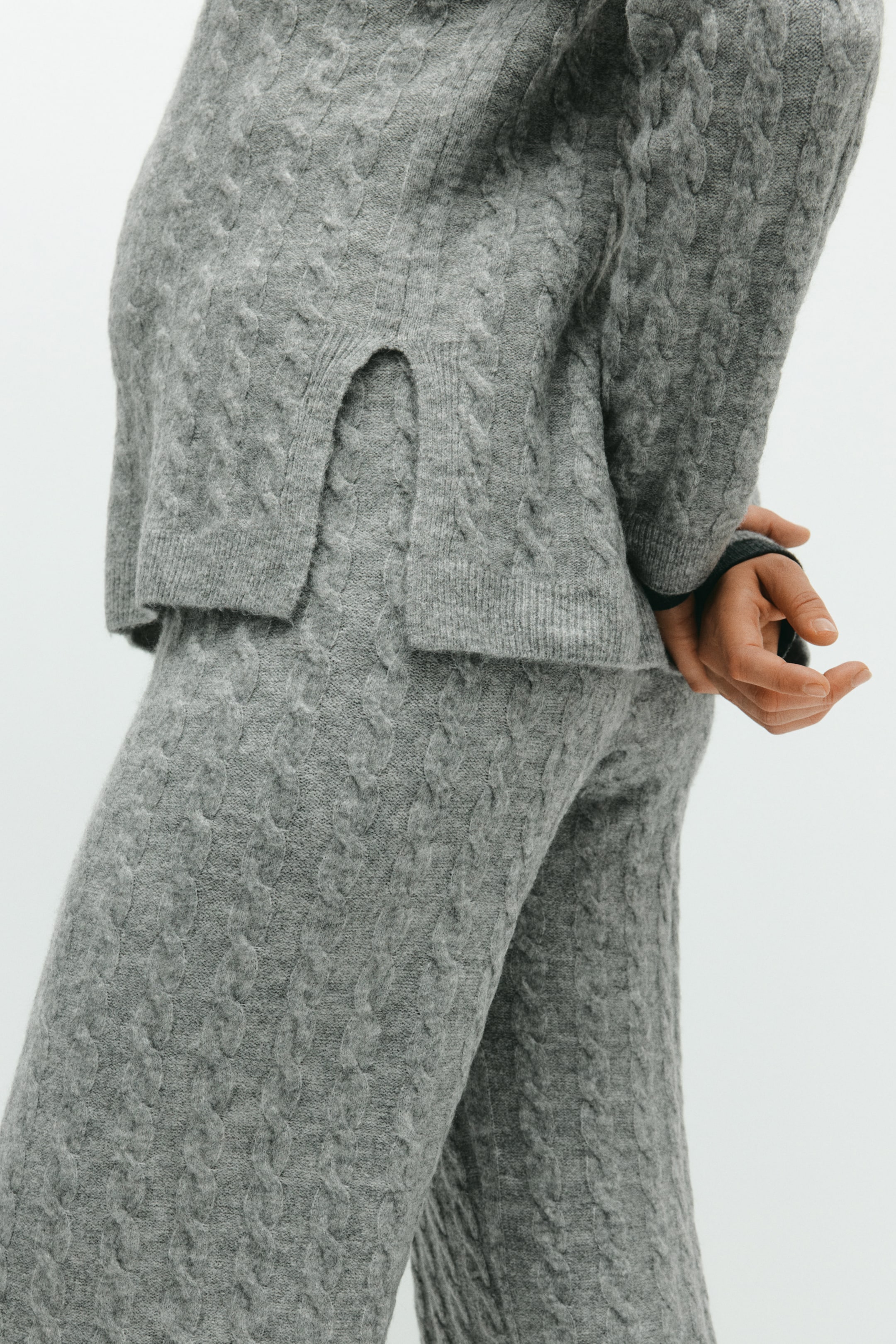 View larger image: A gray cable-knit sweater and coordinating pants are being presented from a side angle. The sweater is featuring a ribbed hem with a side slit, and both pieces display a textured cable-knit pattern.