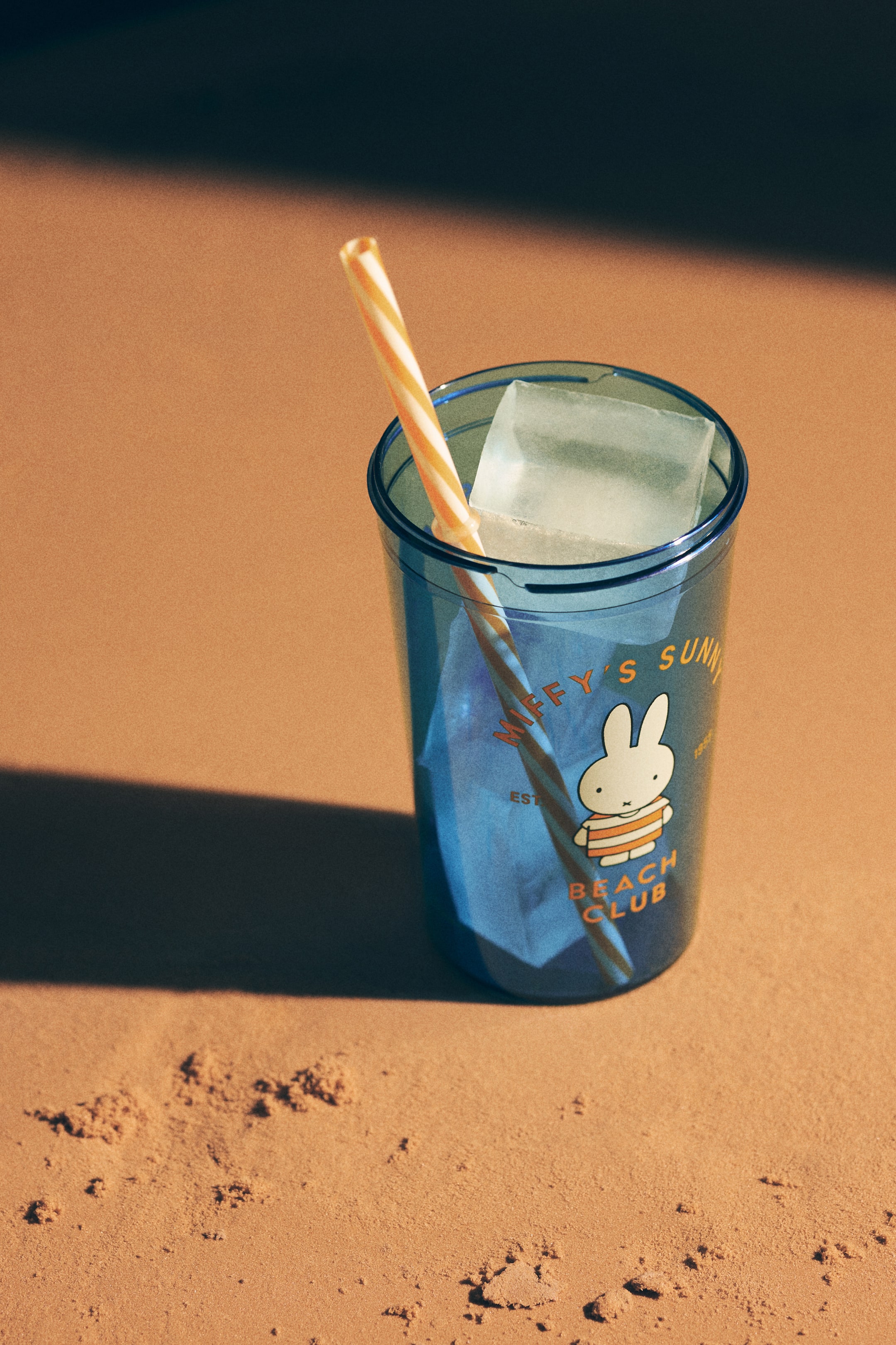 Agrandir l'image: Un gobelet en plastique bleu translucide est posé debout, contenant des glaçons et un liquide clair. Une paille rayée orange et blanche est insérée dans le gobelet, qui présente un graphique orange de Miffy portant une chemise rayée, accompagné des textes "Miffy's Sunny" et "Beach Club".
