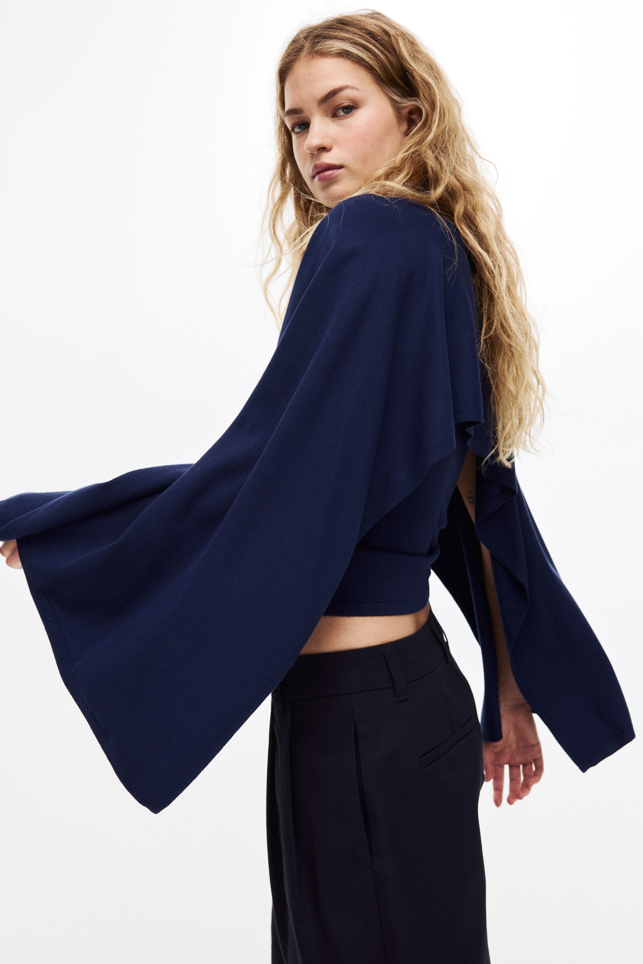 Ampliar la imagen: Se presenta un top corto azul marino, con mangas anchas y fluidas que se abren por la parte inferior. El top envuelve el torso, con su dobladillo por encima de la cintura, revelando unos pantalones negros.