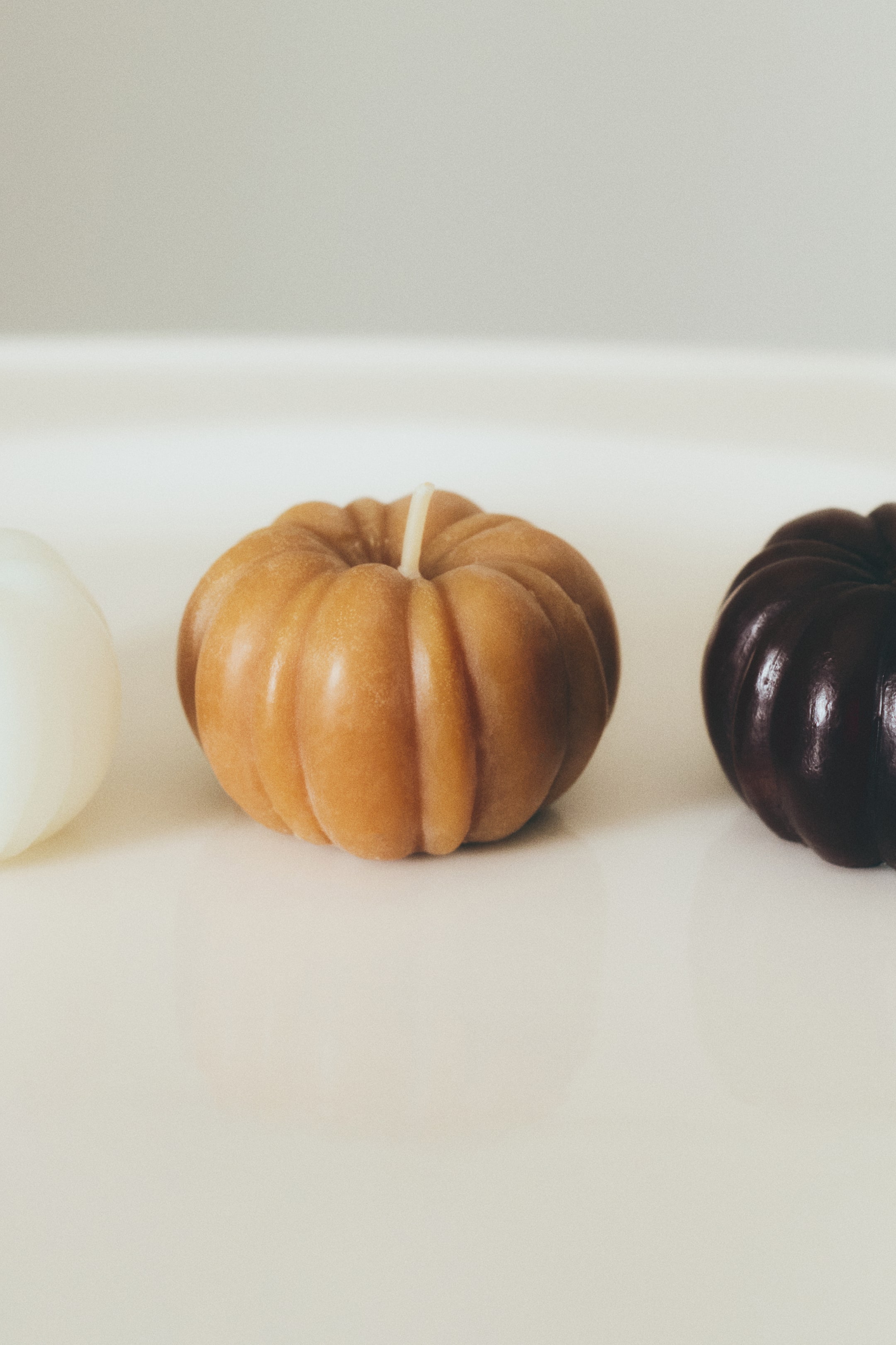 View larger image: Three pumpkin-shaped candles are standing in a row, featuring a central light-brown candle with a ribbed texture and a single wick protruding from the top. A cream-coloured candle is partially visible on the left, and a dark brown candle is partially visible on the right.