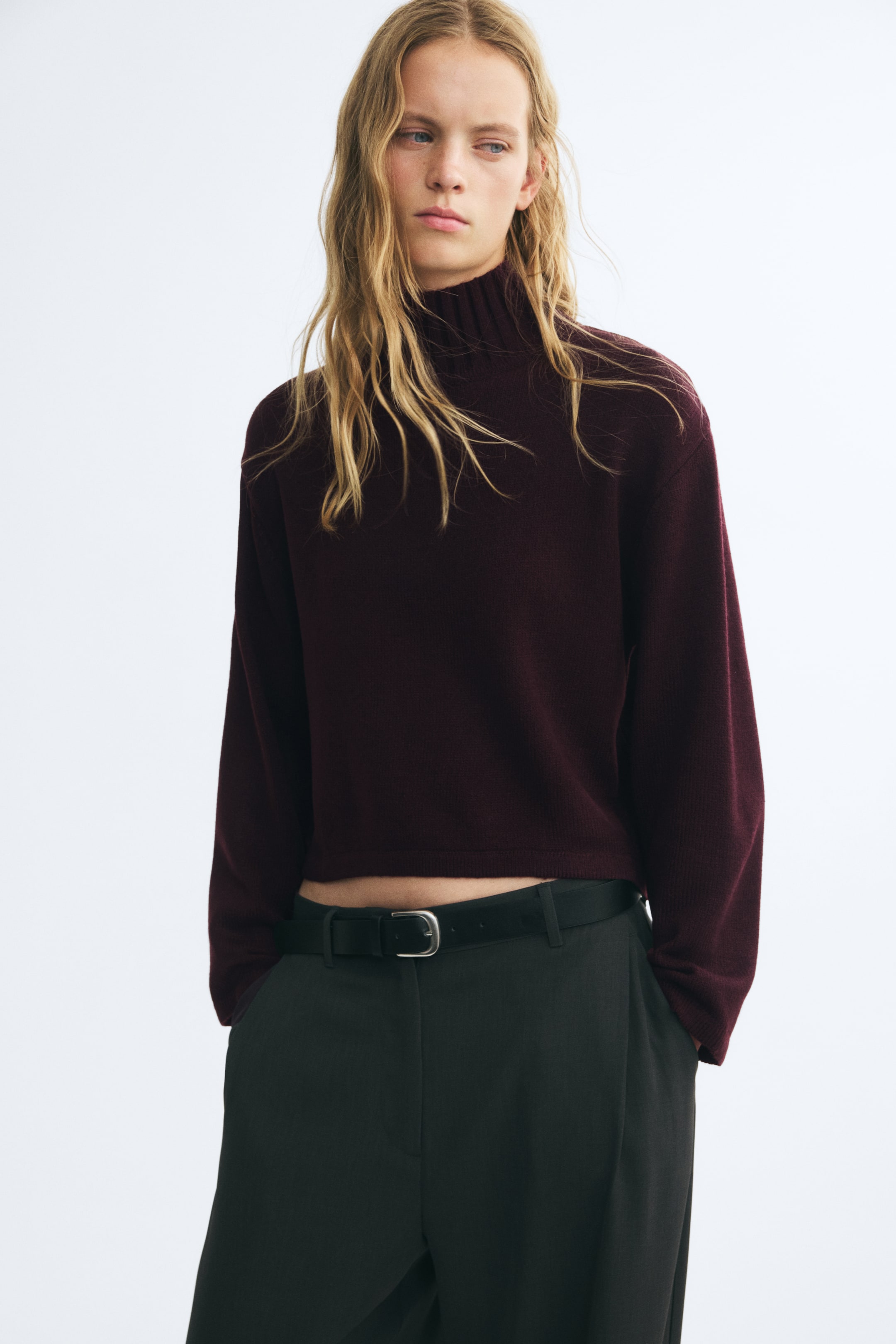 View larger image: A dark burgundy knitted sweater featuring a high ribbed neck, dropped shoulders, and long sleeves is worn by the model. The slightly cropped sweater is styled with dark trousers and a black belt.