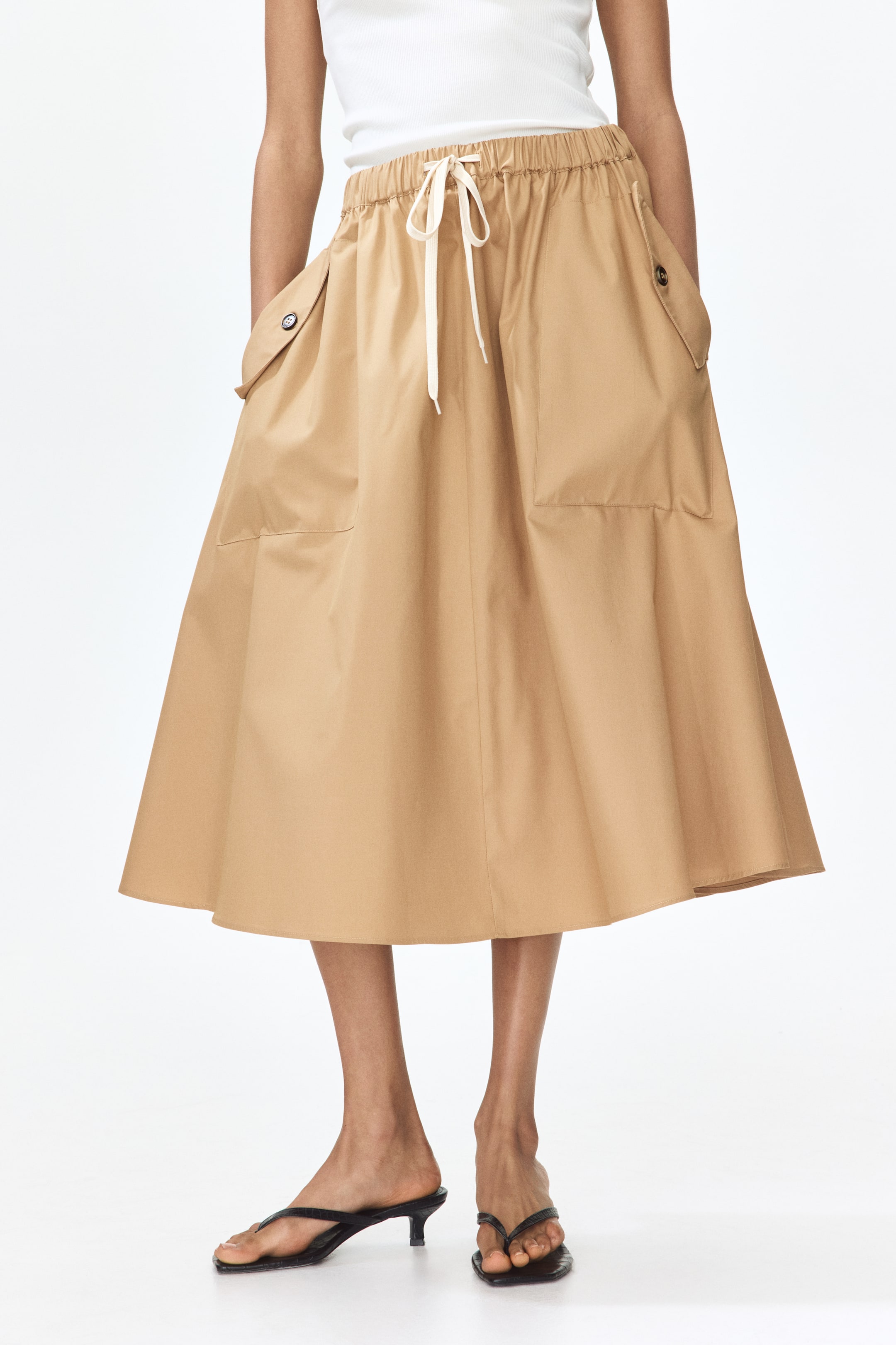 View larger image: A tan A-line midi skirt is being worn, featuring an elasticated drawstring waist with a cream tie. It is displaying two large patch pockets and two smaller flap pockets with button closures on the front.