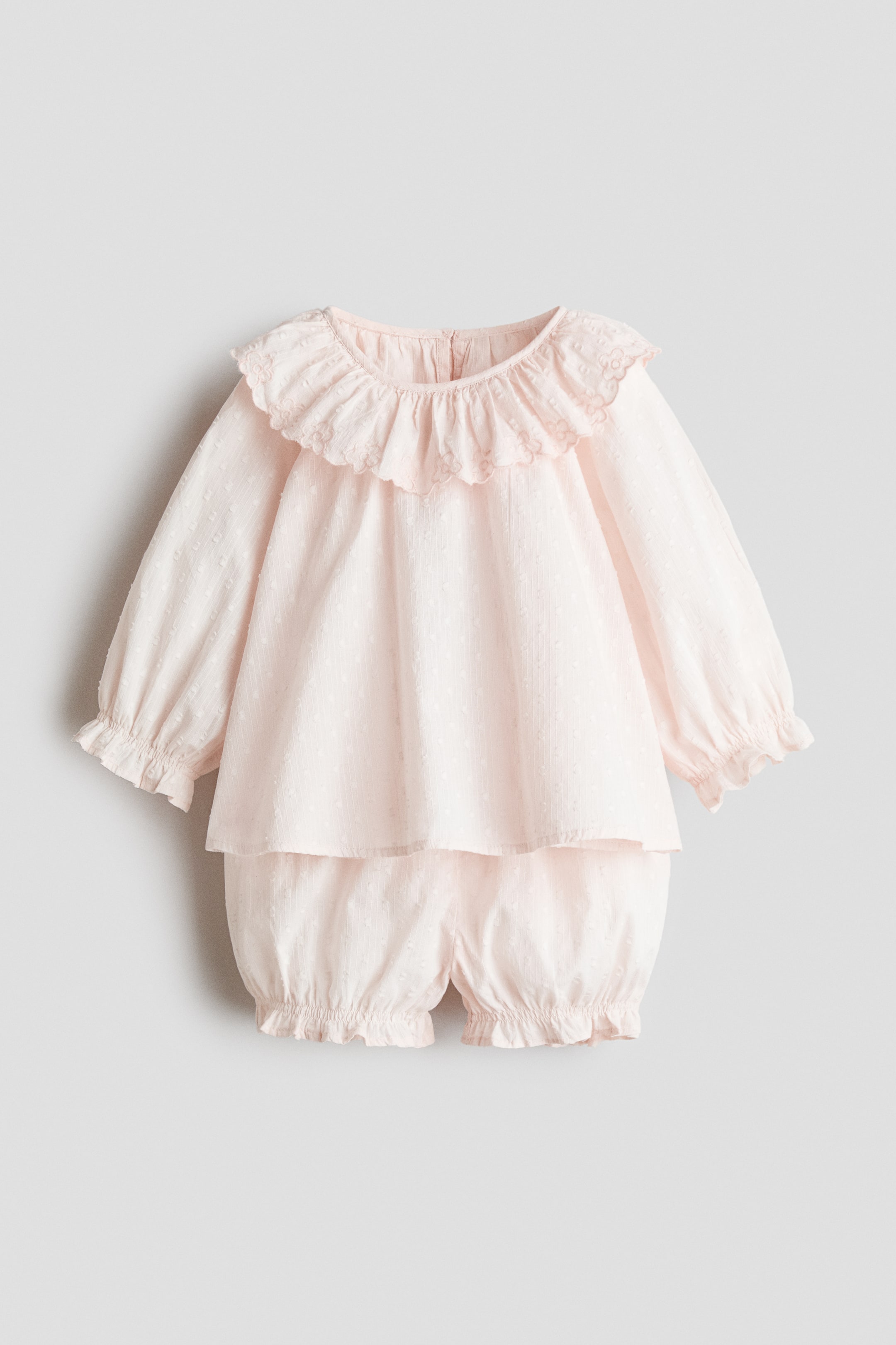 View larger image: A light pink baby outfit is showcased front-facing, featuring a long-sleeved top and bloomer shorts, both made from a textured fabric with small raised dots. The top is designed with a ruffled collar highlighting embroidered detailing and ruffled cuffs, while the shorts are styled with ruffled leg openings.