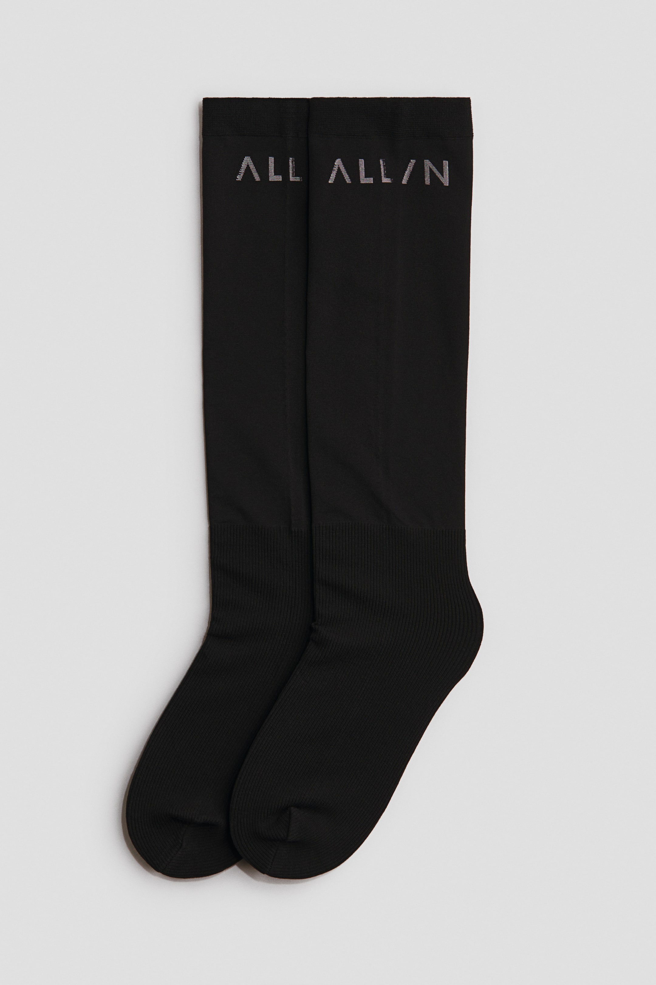 View larger image: A pair of black knee-high riding socks is shown from the front, featuring a fine-knit texture across the main body, ribbed cuffs at the foot, and a jacquard-knit "ALL ALL/N" motif at the top.