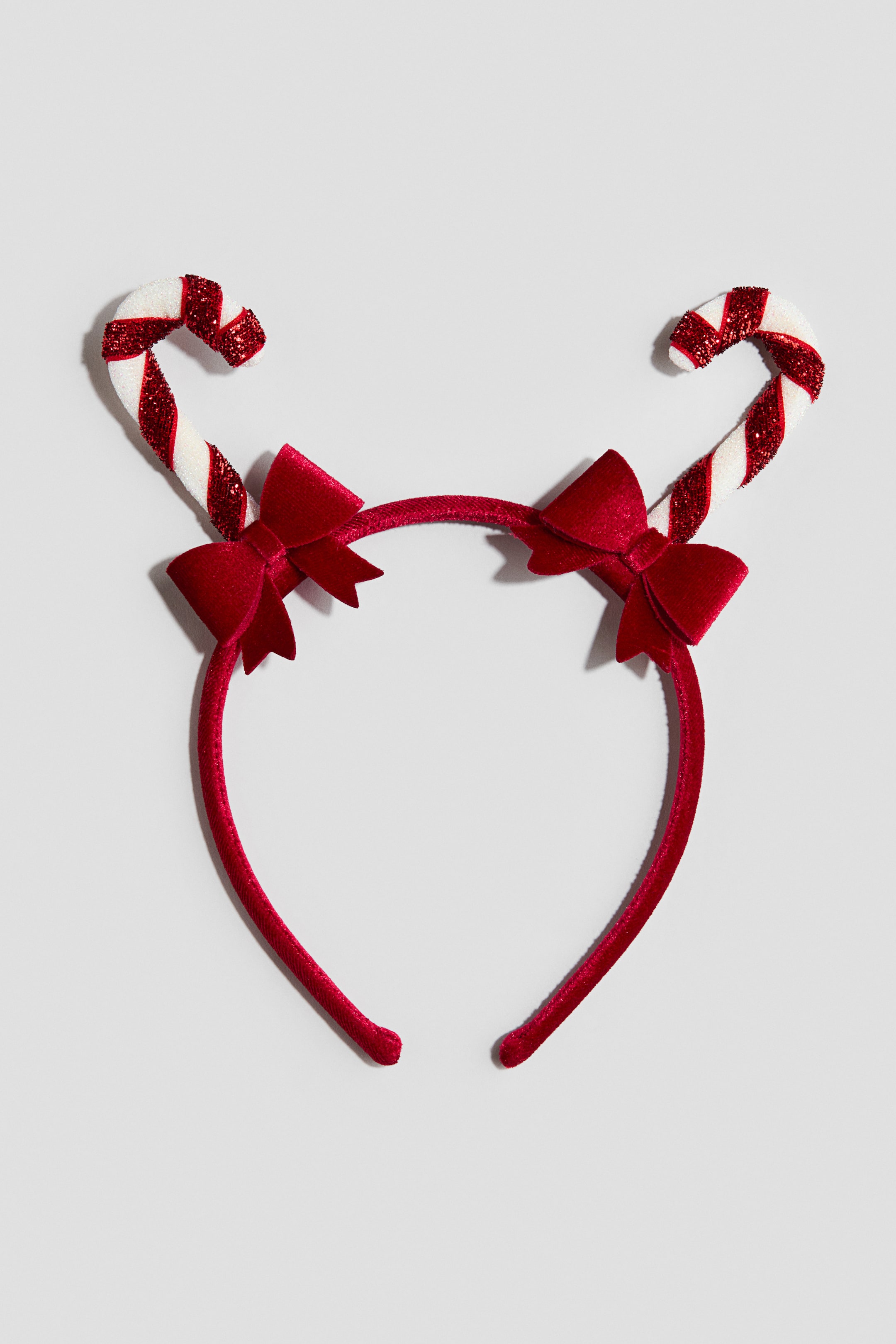 View larger image: A red velvet-covered headband is shown from the front, featuring two candy canes curving outwards from the top. The candy canes are decorated with alternating white and glittery red stripes, with a small red velvet bow attached at the base of each.
