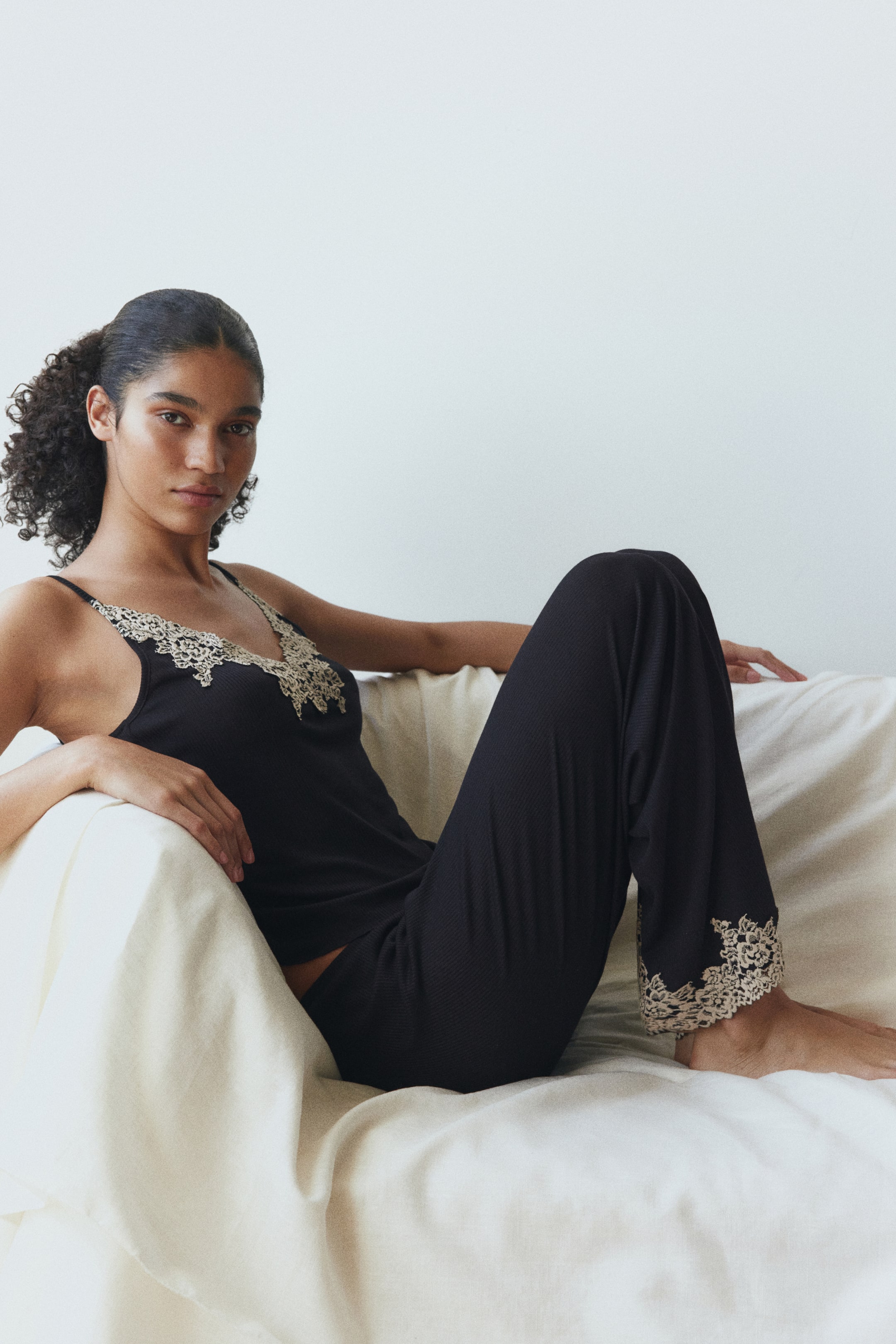 Büyük resmi görüntüle: Model, siyah fitilli bir askılı üst ve uyumlu pijama pantolonu giymektedir. Askılı üstün yakasında açık renkli dantel biye detayları, pijama pantolonunun bilek kısmında ise uyumlu dantel işlemeler yer almaktadır.
