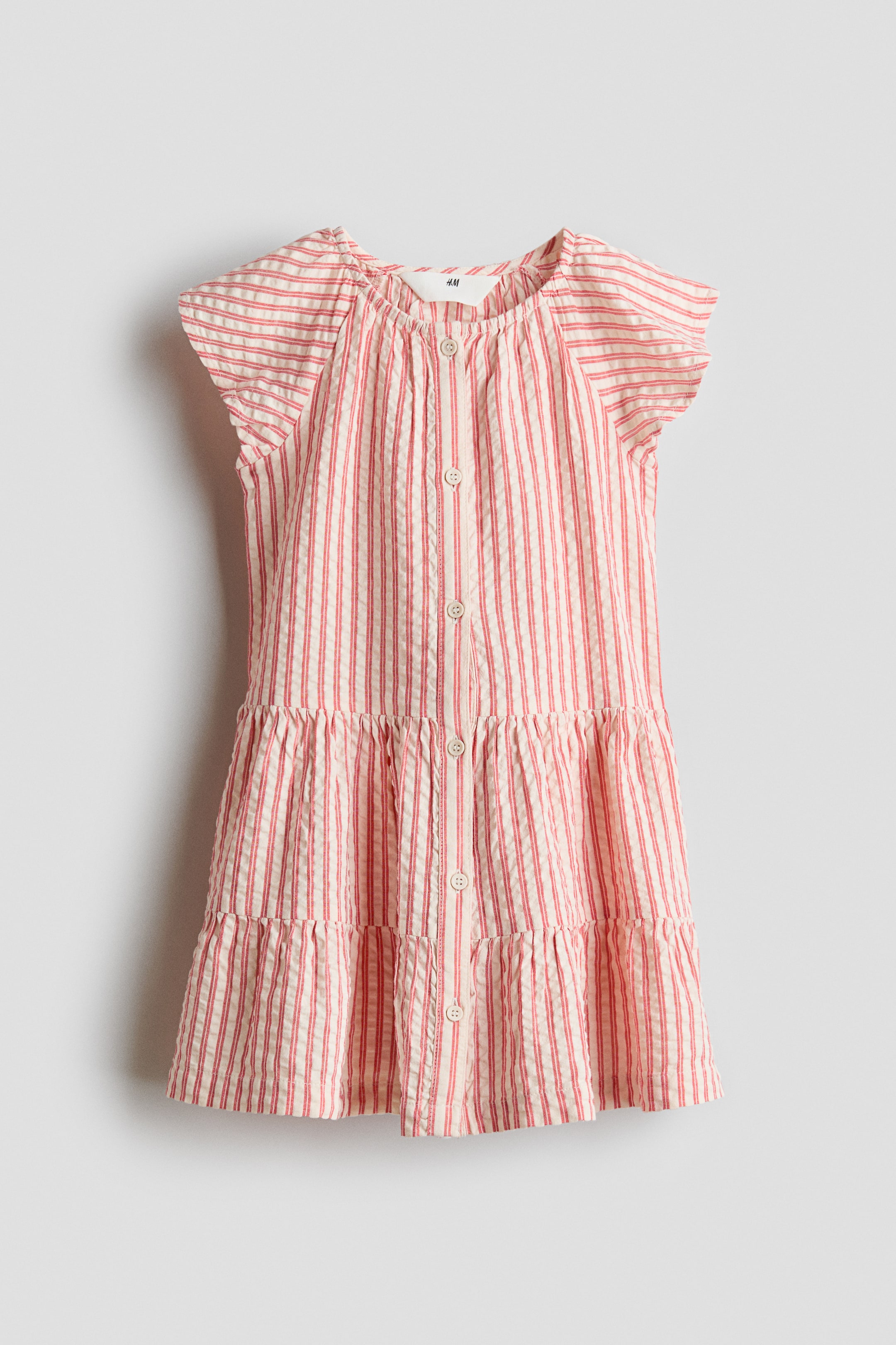 Agrandir l'image: Une robe en seersucker à rayures verticales blanches et rouges est présentée de face, avec un col rond, des manches courtes à épaulettes froncées, et une patte de boutonnage complète sur le devant. La robe se termine par deux volants superposés, formant une jupe ample.