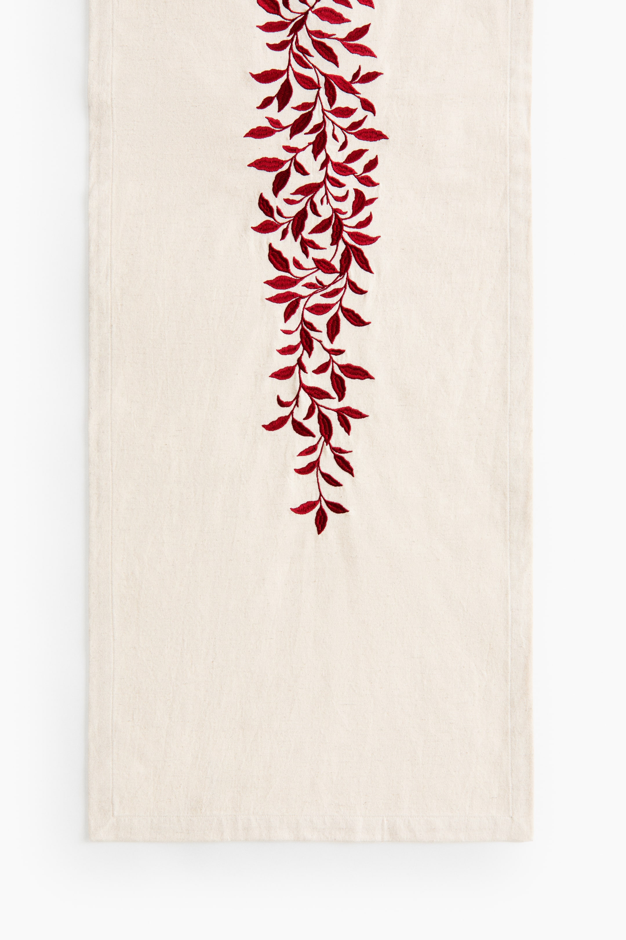 View larger image: A cream-coloured fabric table runner is shown from the front, featuring a vertical embroidered vine of dark red leaves cascading down the centre. The runner displays a woven texture with clean stitched hems along its borders.