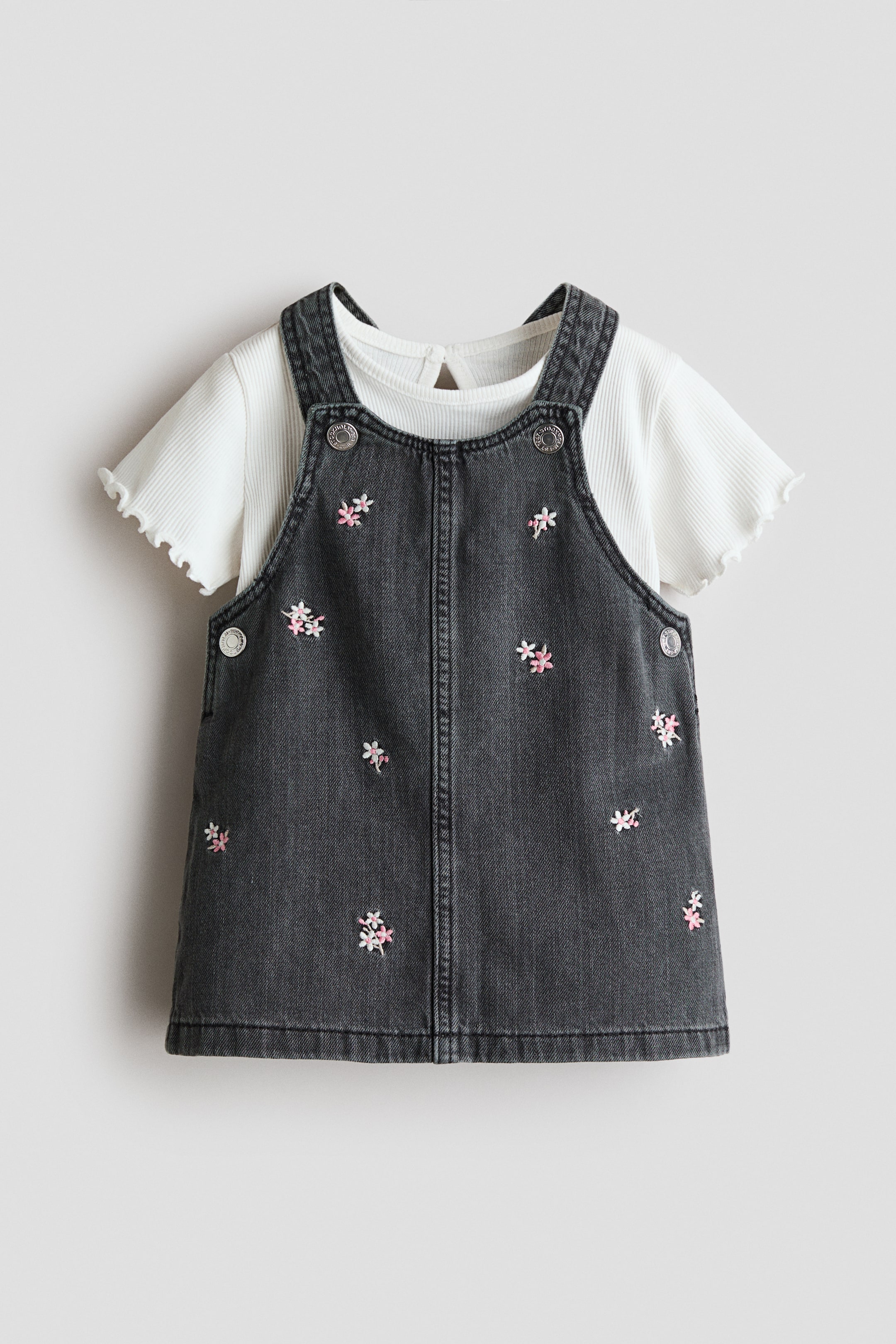 Grösseres Bild ansehen: Ein gewaschenes schwarzes Denim-Latzkleid zeigt rosa und weiße gestickte Blumenmotive, silberfarbene Knöpfe am Latz und seitliche Verschlüsse, von vorne betrachtet. Es ist über einem weißen gerippten T-Shirt mit rundem Ausschnitt, kurzen Ärmeln mit Rüschenbündchen und einem Schlüssellochausschnitt im Nacken geschichtet.