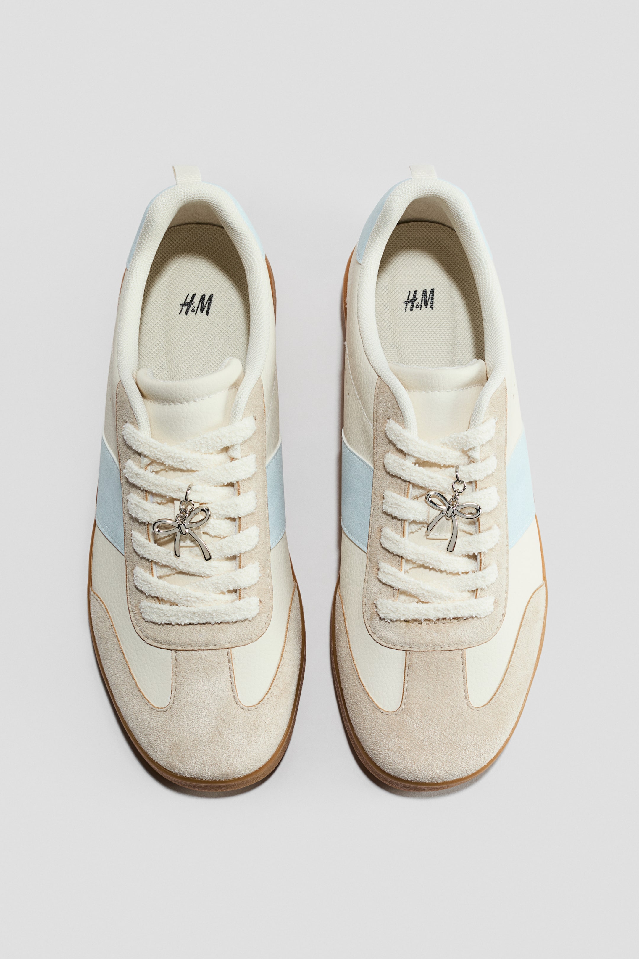 Näytä suurempi kuva: Lenkkareissa on kermaiset päällisosat, beiget mokkanahkaiset päällysteet ja vaaleansiniset sivuraidat. Jokaisessa lenkkarissa on hopeansävyinen rusettiriipus, joka on pujotettu sen kermaisiin, teksturoituihin nauhoihin.