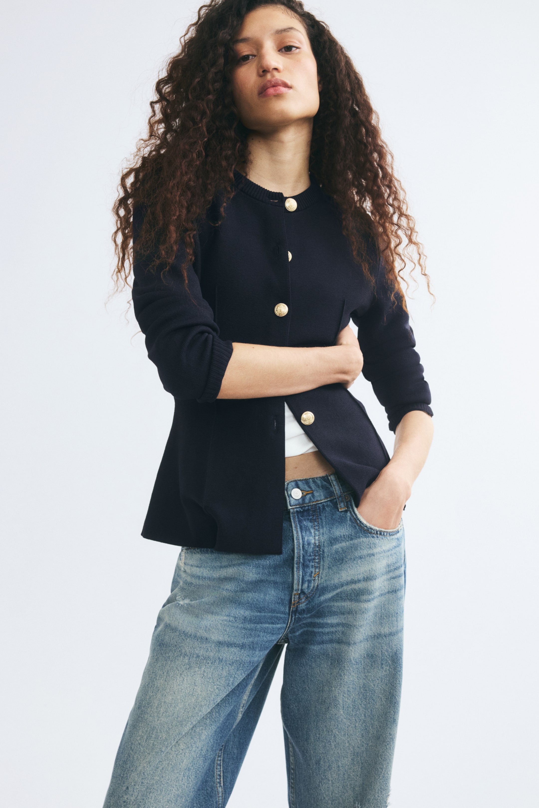 Voir une image plus grande: Un cardigan ajusté en maille fine bleu marine foncé, doté d’un col rond côtelé et de boutons métalliques dorés, est porté par le mannequin. Le cardigan est stylisé avec le bas partiellement déboutonné, laissant entrevoir un haut blanc en dessous, et il est assorti à un jean bleu.