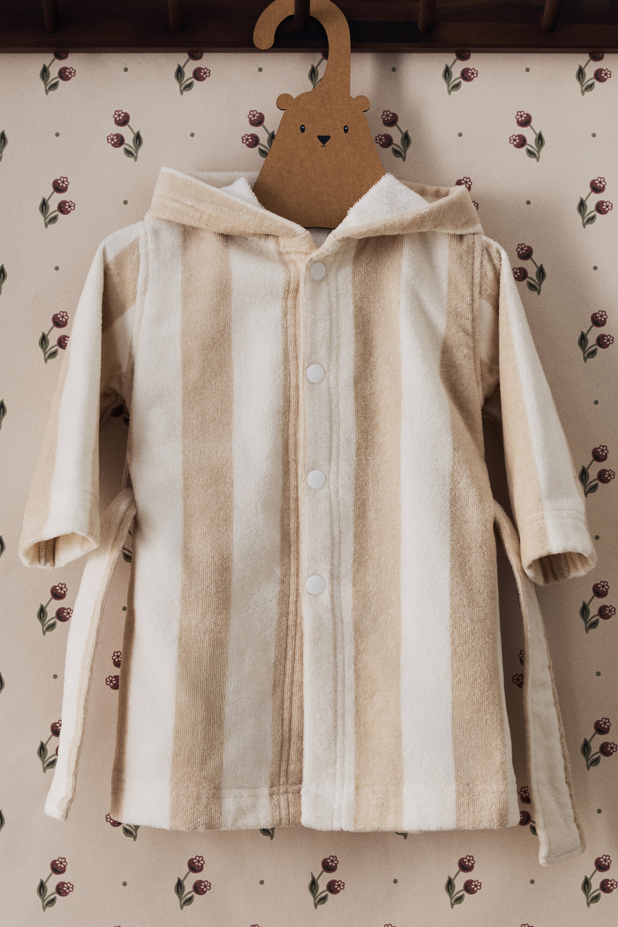 Nagyobb kép megtekintése: Egy kapucnis gyerek köntös elölnézete látható, amely bézs és törtfehér frottír anyagból készült, váltakozó függőleges csíkokkal. Hosszú ujjakkal, rögzített kapucnival és középen elhelyezkedő elülső pánttal rendelkezik, amelyet több fehér gomb rögzít, és az oldalakról szövetkötők nyúlnak ki.