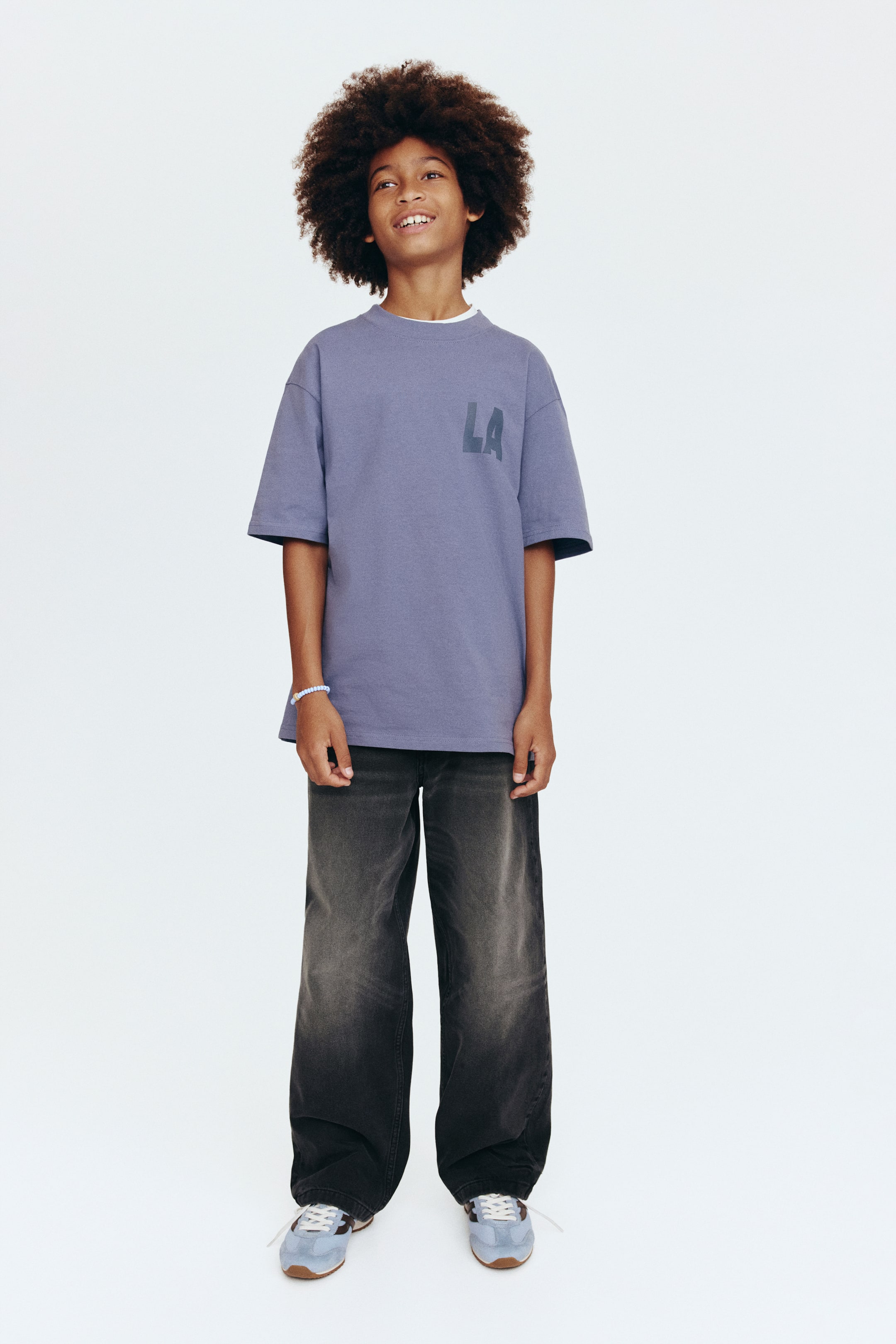 Voir une image plus grande: Un t-shirt surdimensionné de couleur violet poussiéreux, avec un col rond et un motif "LA" sur la poitrine gauche, est porté en superposition sur un col ras du cou blanc. Il est associé à un jean noir délavé à jambes larges et des baskets bleu clair et marron avec des lacets blancs.