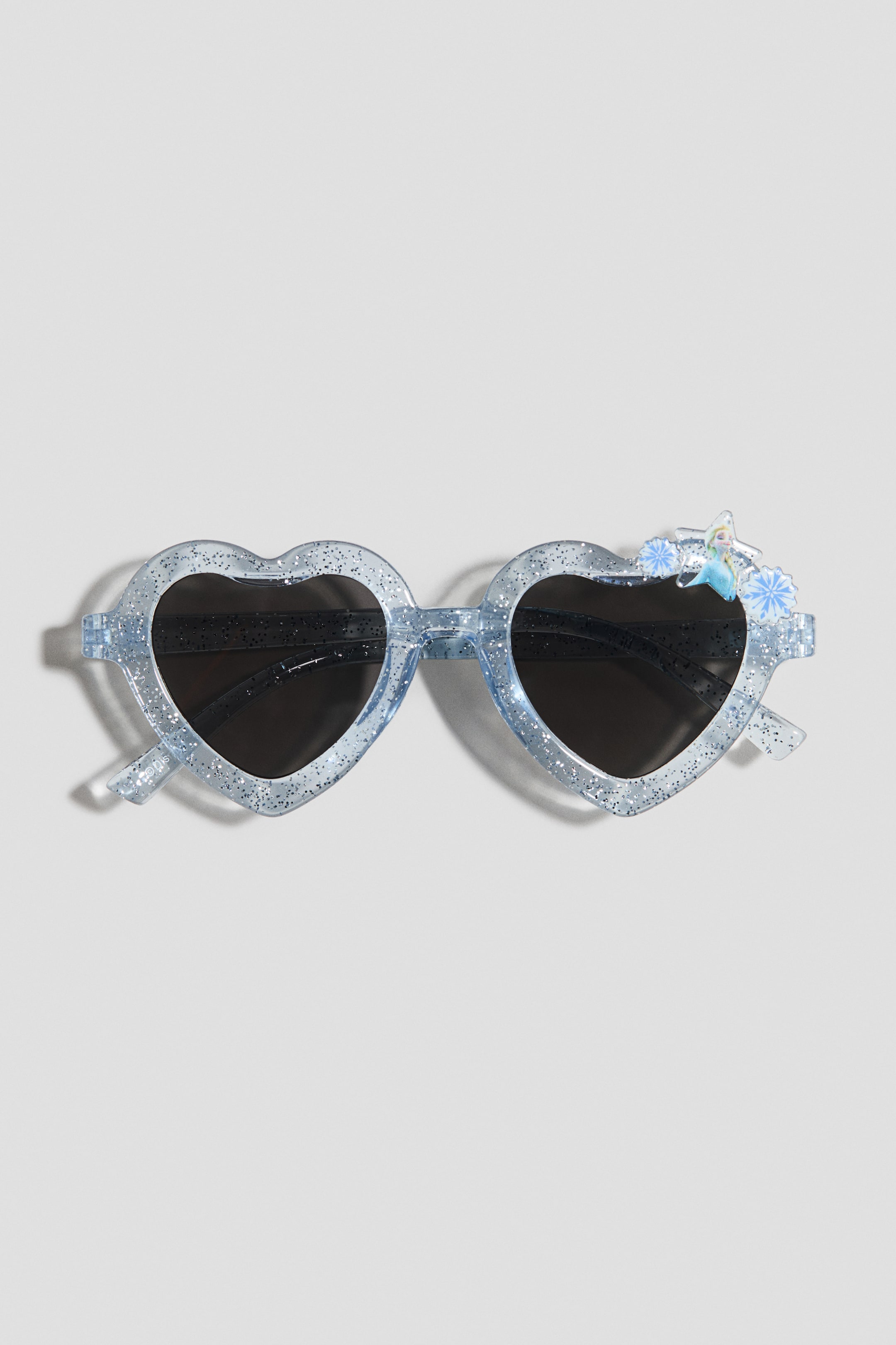 Ampliar la imagen: Las gafas de sol presentan monturas en forma de corazón, translúcidas y de color azul claro con brillo, vistas de frente. Lentes oscuros están montados en ellas, y una figura de personaje rodeada de copos de nieve azules adorna la esquina superior derecha.