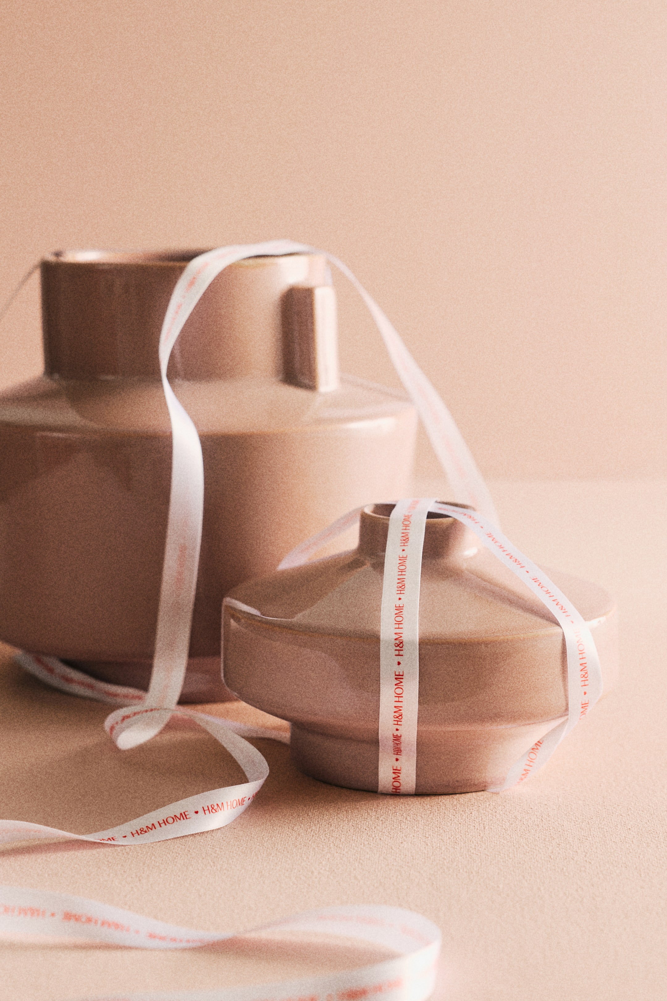 이미지 확대 보기: 키가 더 큰 토프 톤의 세라믹 화병은 직사각형 목과 더 넓은 받침을 드러내고 있으며, 더 짧고 넓은 토프 톤의 세라믹 화병은 원반 모양의 윗부분과 좁은 입구를 특징으로 합니다. 'H&M HOME' 로고가 들어간 흰색 리본이 키가 더 큰 화병의 목에 걸쳐 있고, 더 짧은 화병의 몸체를 감싸고 있습니다.