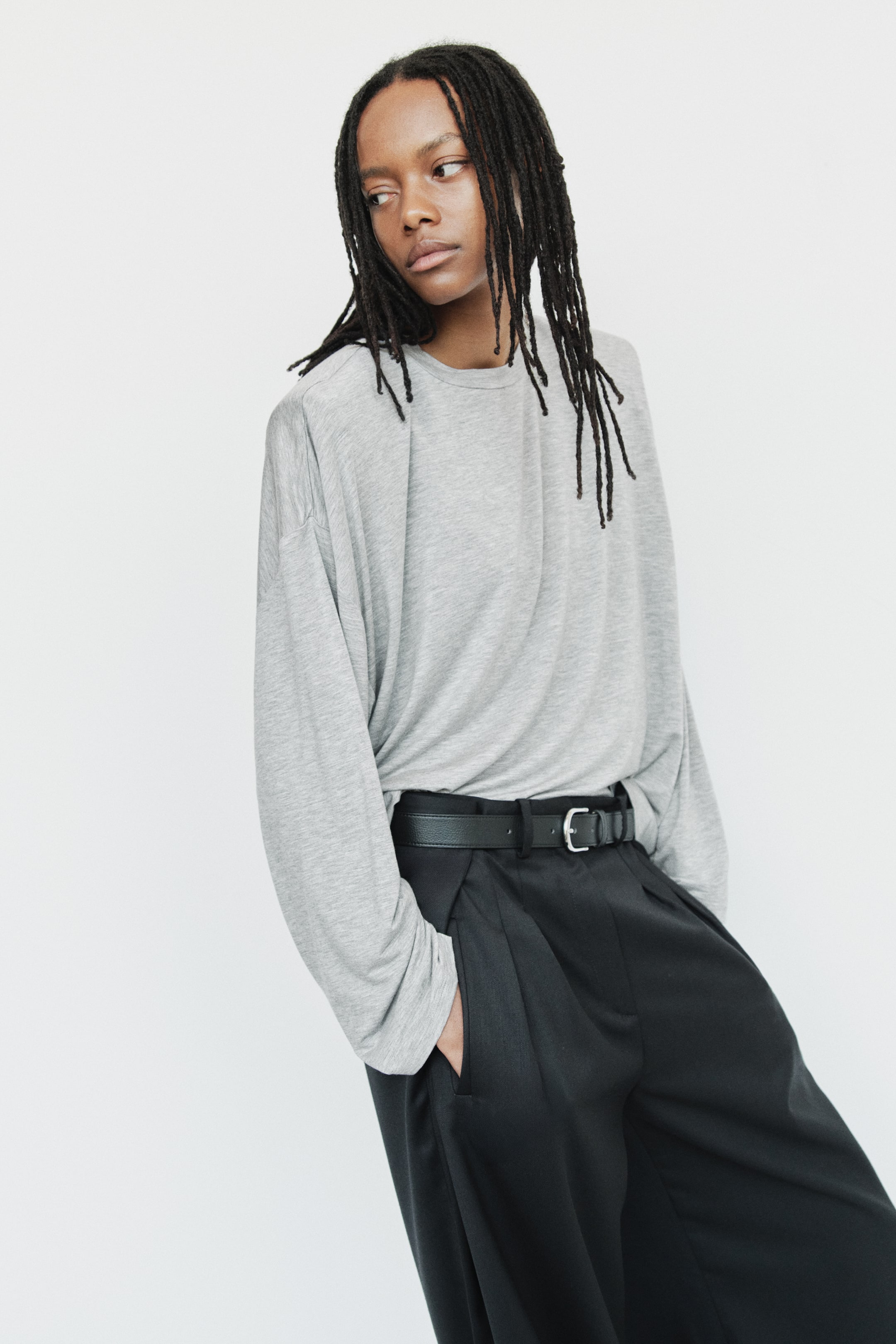 View larger image: A loose-fit top in light grey is presented from the front, featuring a round neckline with a narrow trim, dropped shoulders, and long sleeves. It is partially tucked into black trousers.