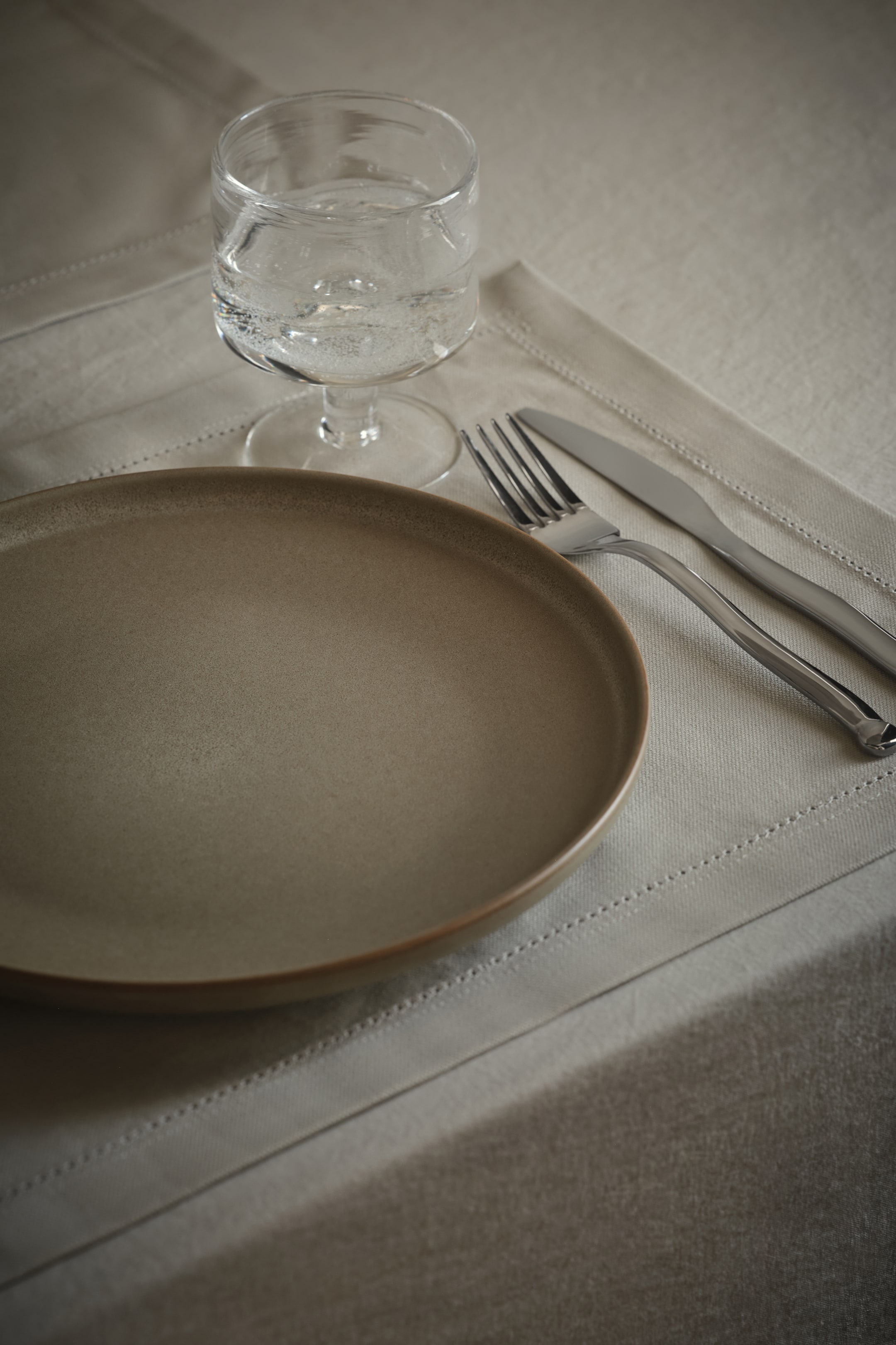 Visualizza immagine più grande: Una mise en place viene presentata, caratterizzata da un piatto rotondo di colore grigio-marrone screziato con un bordo rialzato, un bicchiere trasparente con stelo riempito d'acqua e una forchetta e un coltello color argento lucido. Questi sono posizionati su una tovaglietta color beige chiaro con bordi cuciti.