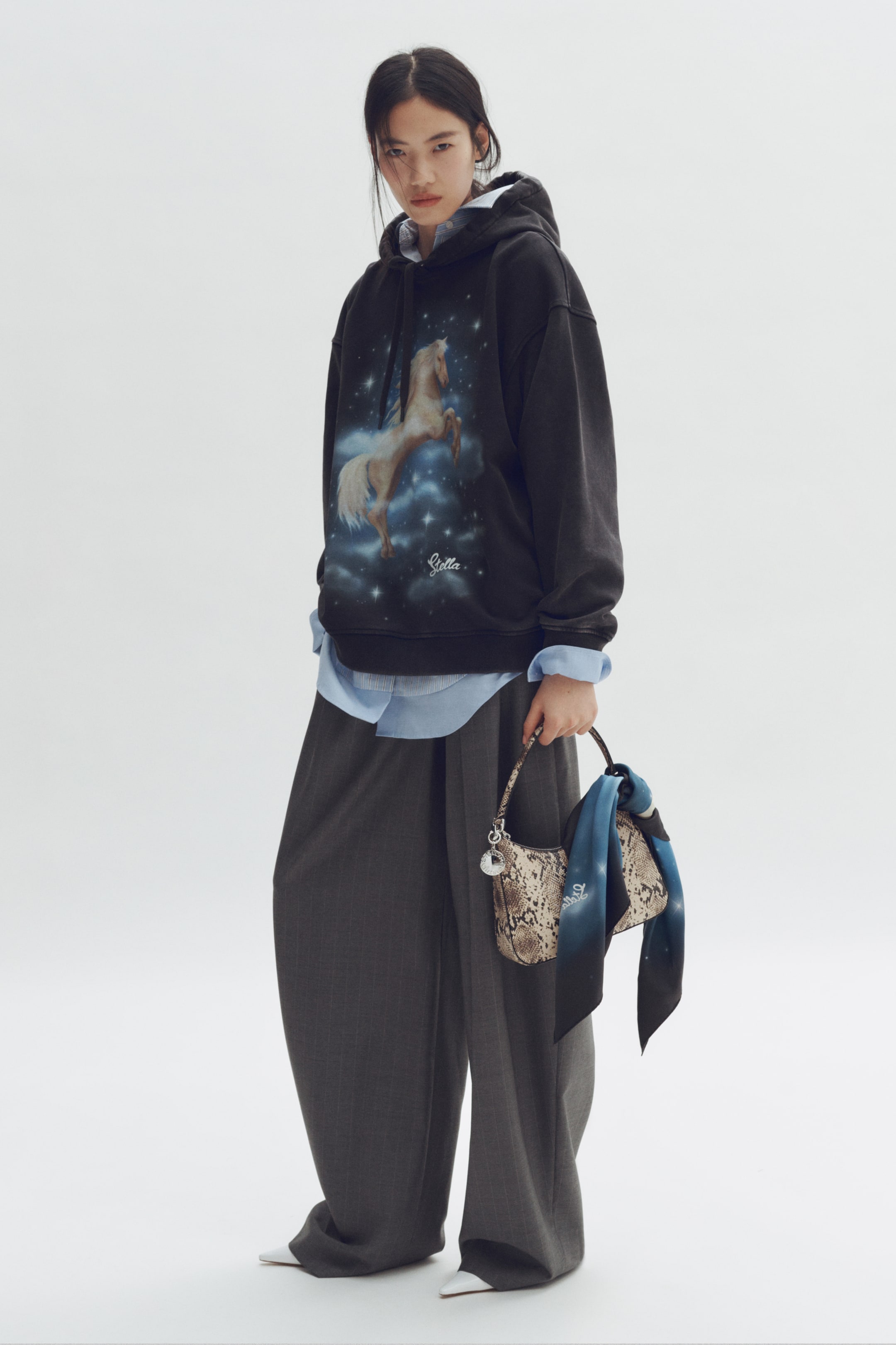 Xem ảnh lớn hơn: Áo hoodie đen dáng rộng với hình in đồ họa của một con ngựa màu kem đang dựng đứng trong bầu trời xanh đầy sao, với chữ "Stella" bên dưới. Áo có thiết kế cổ tay và gấu áo bo gân, được phối cùng áo sơ mi cổ bẻ màu xanh nhạt.