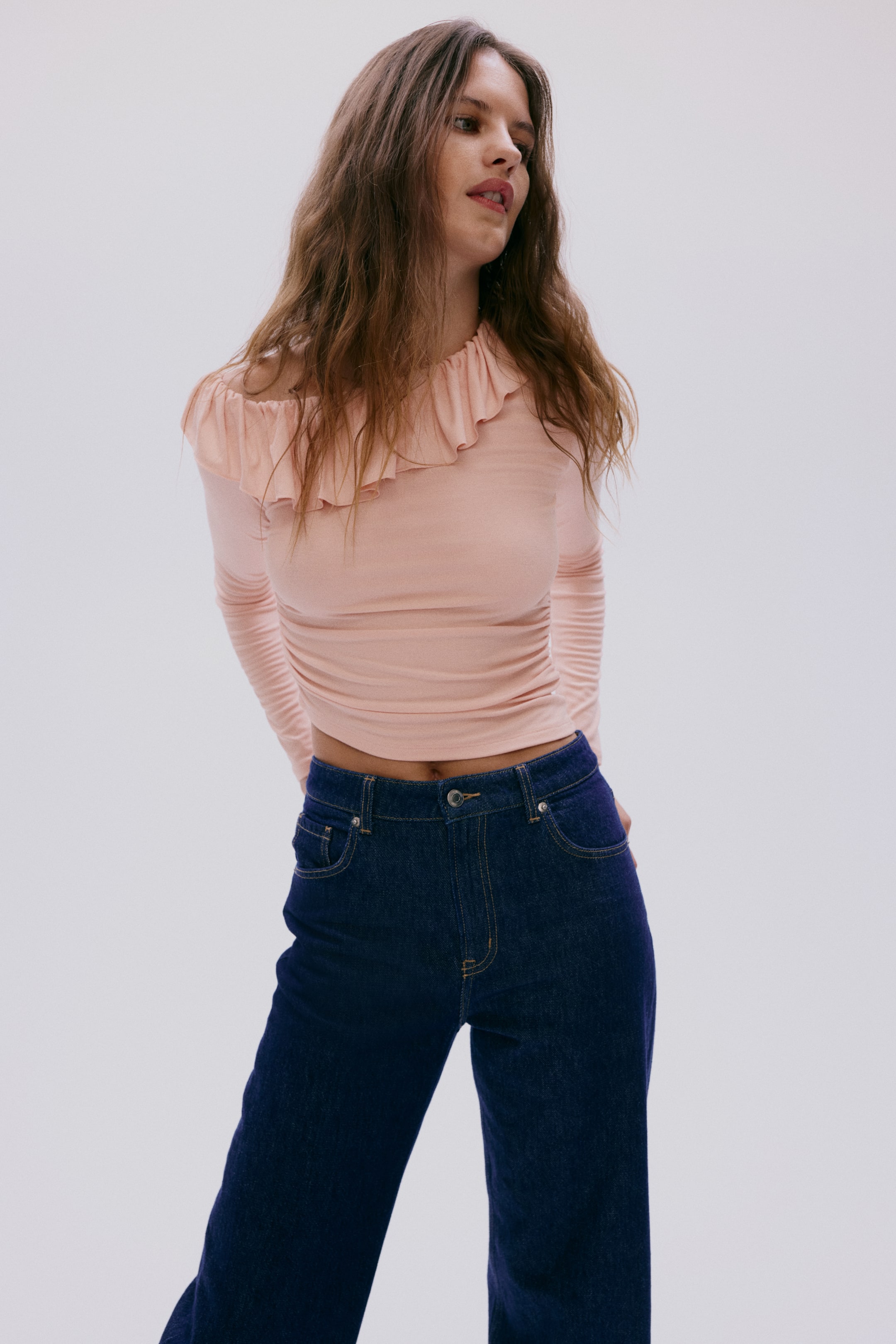 View larger image: A light pink long-sleeved top na may ruffled, off-the-shoulder neckline na nag-eexpose sa kanang balikat, cropped hem, at subtle ruching sa sleeves at katawan. Ito ay isinusuot kasama ng dark blue high-waisted jeans na may front pockets at yellow contrast stitching.