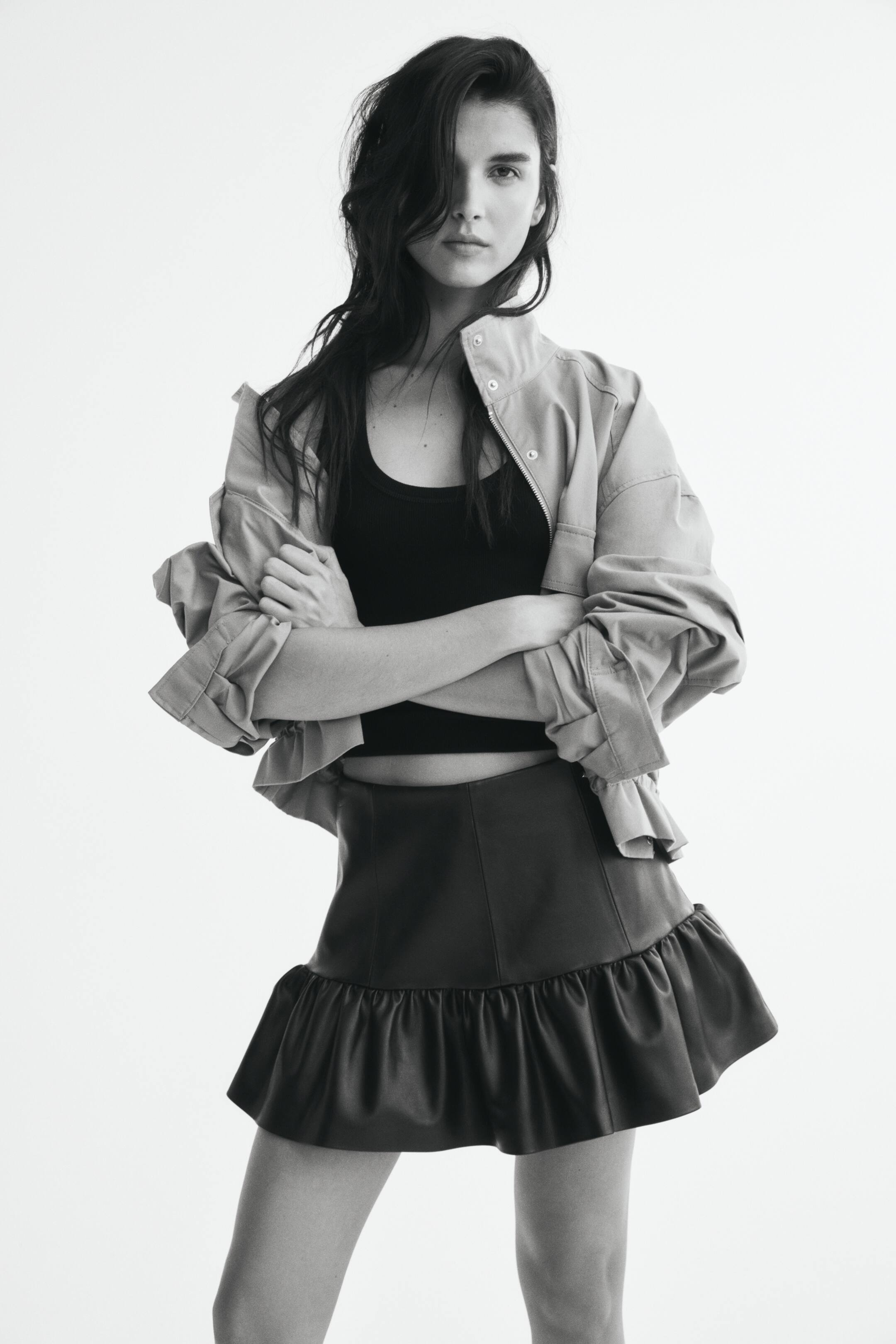 View larger image: The model is wearing a short skirt in coated fabric, featuring a visible vertical seam down the front and a wide, gathered ruffle tier at the hem. The skirt is styled with a fitted cropped top and a light jacket draped off the shoulders.