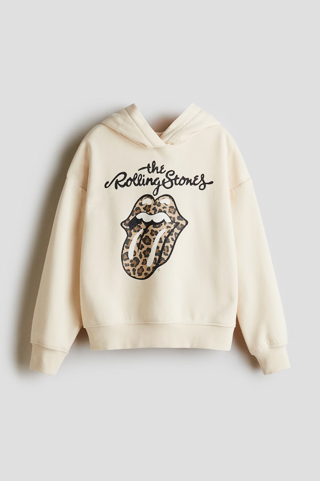 Un chandail à capuchon de couleur crème affiche son DEVANT, mettant en valeur un grand imprimé graphique avec le texte noir 'the Rolling Stones' au-dessus d'un motif de langue et lèvres en imprimé léopard. Ce chandail à capuchon en tissu molletonné présente une capuche doublée avec un devant croisé, des épaules tombantes et des bordures côtelées aux poignets et à l'ourlet. - 1