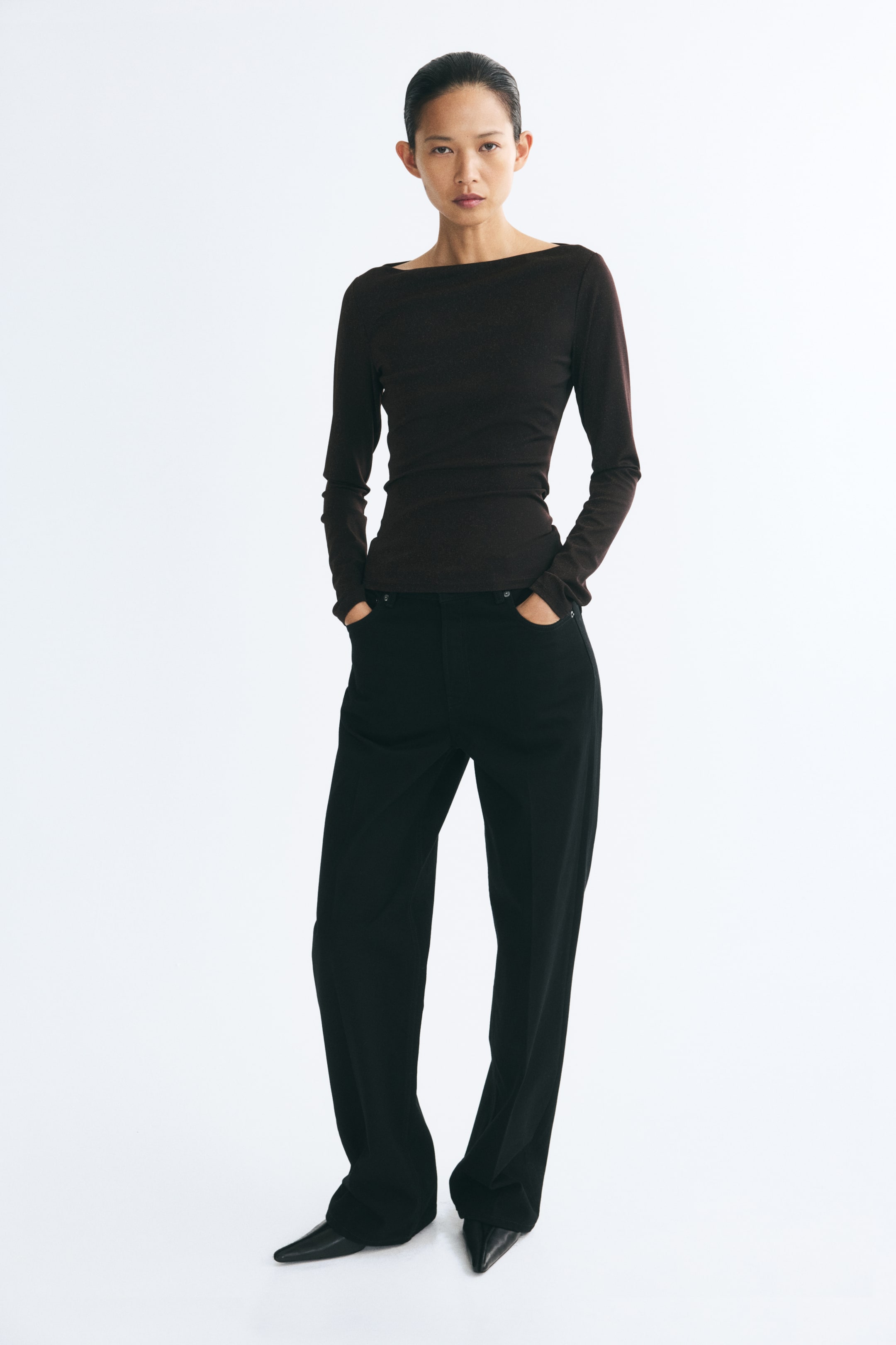 Größeres Bild ansehen: Ein dunkelbraunes, figurbetontes Langarm-Top mit weitem U-Ausschnitt wird vom Model getragen. Das Oberteil ist mit einer schwarzen Wide-Leg-Hose kombiniert, wobei das Model die Hände in den vorderen Taschen ablegt.