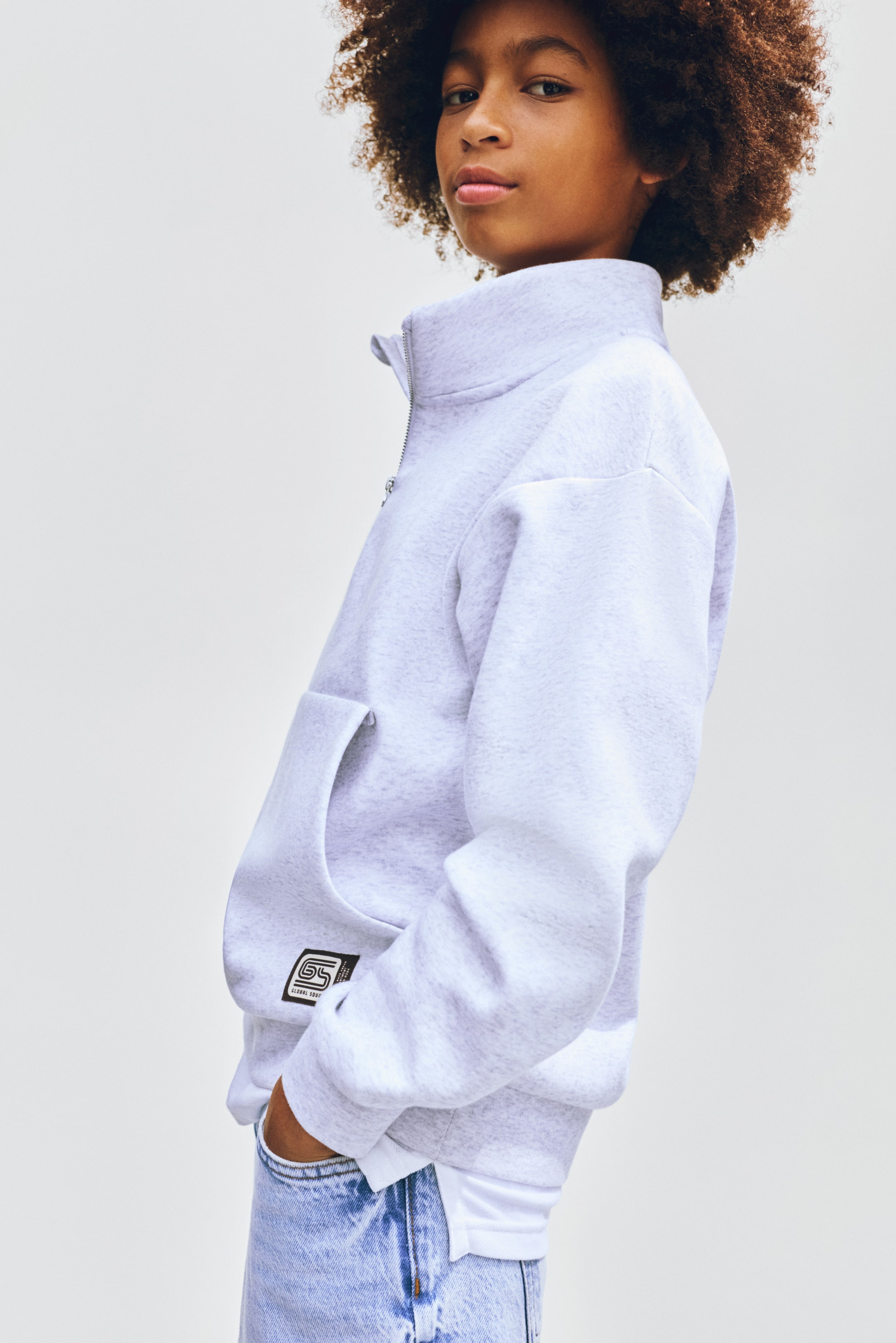 View larger image: The light grey quarter-zip sweatshirt showcases a stand-up collar with a silver zipper and long sleeves with ribbed cuffs. Presented from a slight side angle, it highlights the structure of the stand-up collar and the depth of the kangaroo pocket. It features a front kangaroo pocket adorned with a black and white logo patch, with a white t-shirt subtly visible underneath.