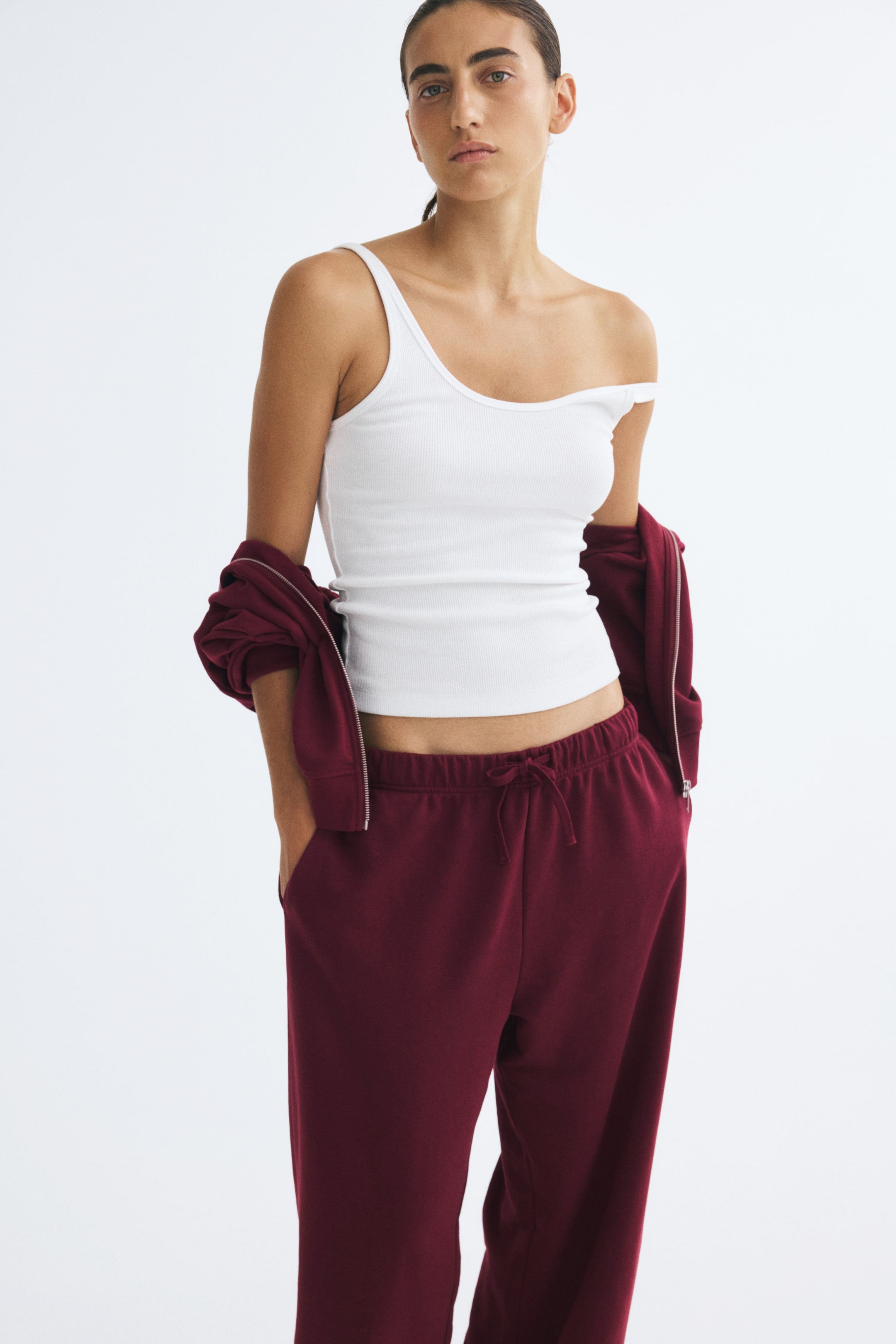View larger image: Track pants in burgundy sweat fabric with an elasticized drawstring waist are worn by the model, with hands placed in the side pockets. The straight-leg pants are styled with a white ribbed tank top and a matching burgundy zip-up hoodie draped over the arms.