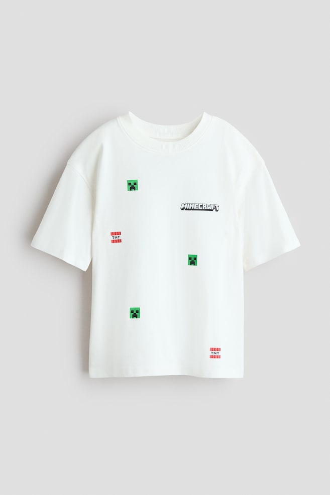 Ein übergroßes, cremefarbenes T-Shirt wird von vorne gezeigt, mit einem gerippten Rundhalsausschnitt und überschnittenen Schultern. Das T-Shirt zeigt ein schwarz-weißes Minecraft-Logo sowie verstreute, verpixelte grüne Creeper-Gesichter und rote TNT-Blöcke auf der Vorderseite. - 1