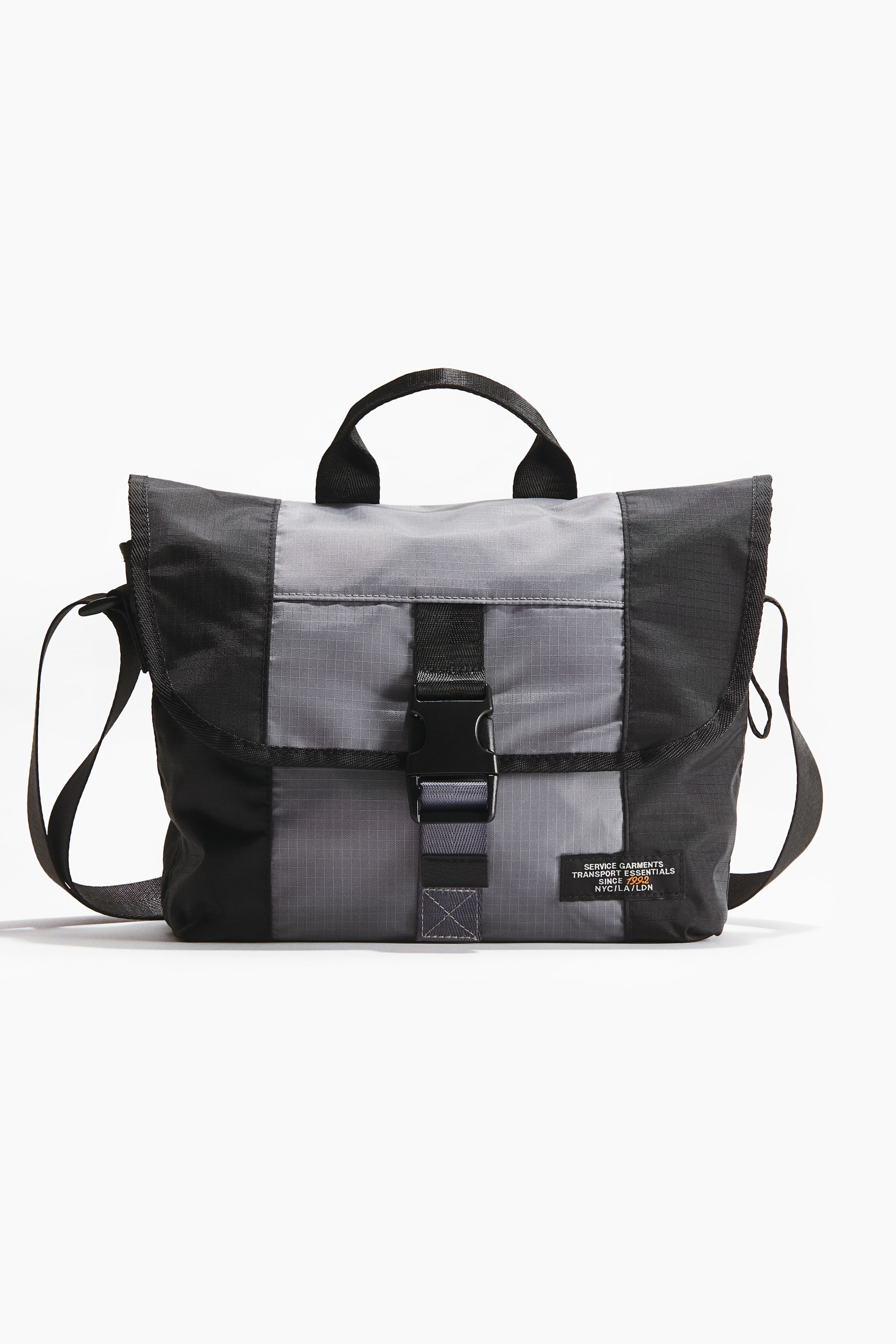 Agrandir l'image: Un sac bandoulière bicolore gris et noir est présenté de face, mettant en avant un tissu tissé durable avec un motif en grille sur les sections grises. Il est doté d'une poignée supérieure noire, d'une bandoulière noire ajustable et d'un rabat à l'avant sécurisé par une sangle noire avec une fermeture à boucle.