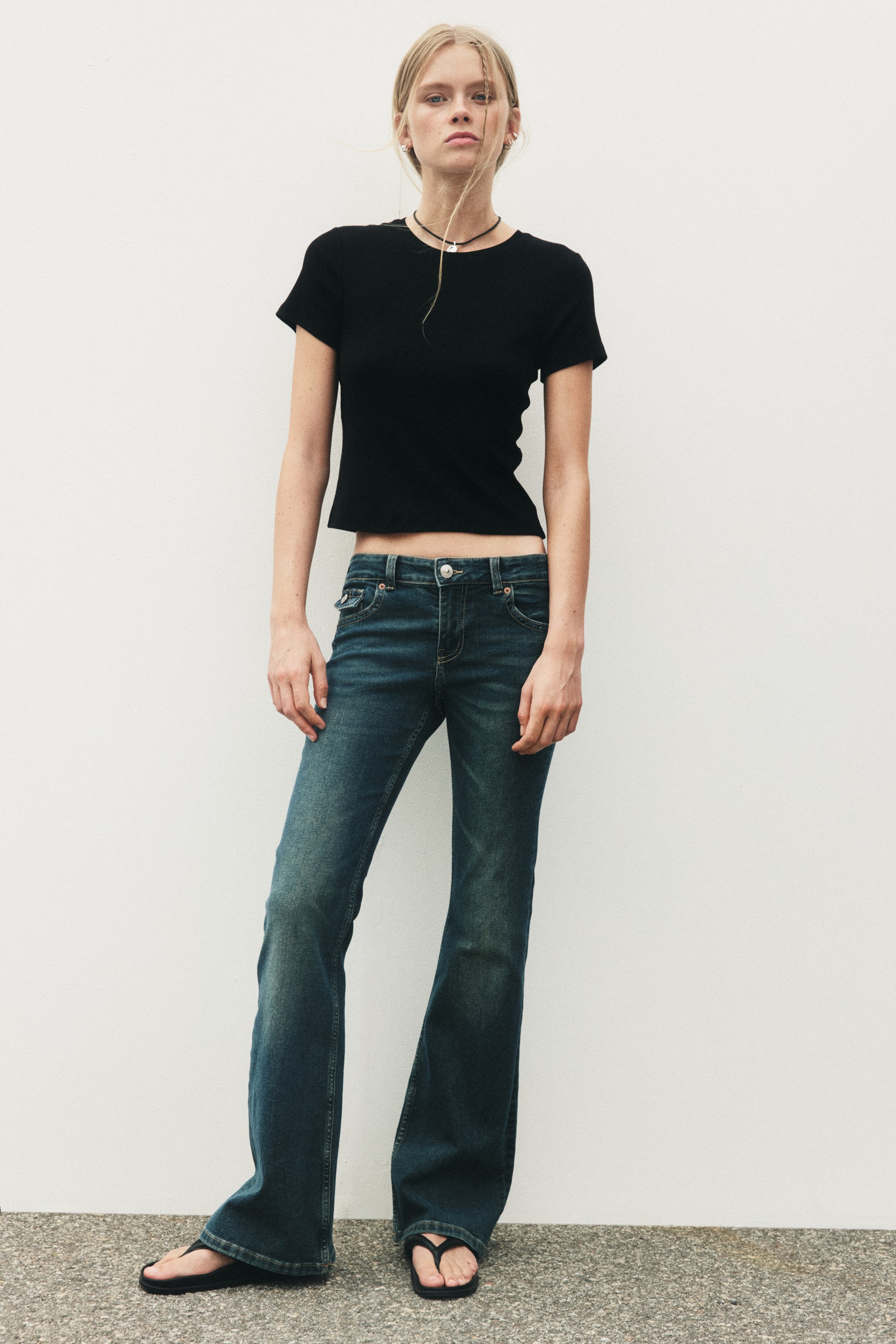 Grösseres Bild ansehen: Der Model trägt dunkelblaue Denim-Jeans mit tiefer Taille, Knopf- und Reissverschluss, Fronttaschen und einer Münztasche. Die Jeans sind am Oberschenkel schmal geschnitten und werden vom Knie bis zum Saum ausgestellt, wobei der Saum in regulärer Länge auf den Füssen des Models aufliegt.