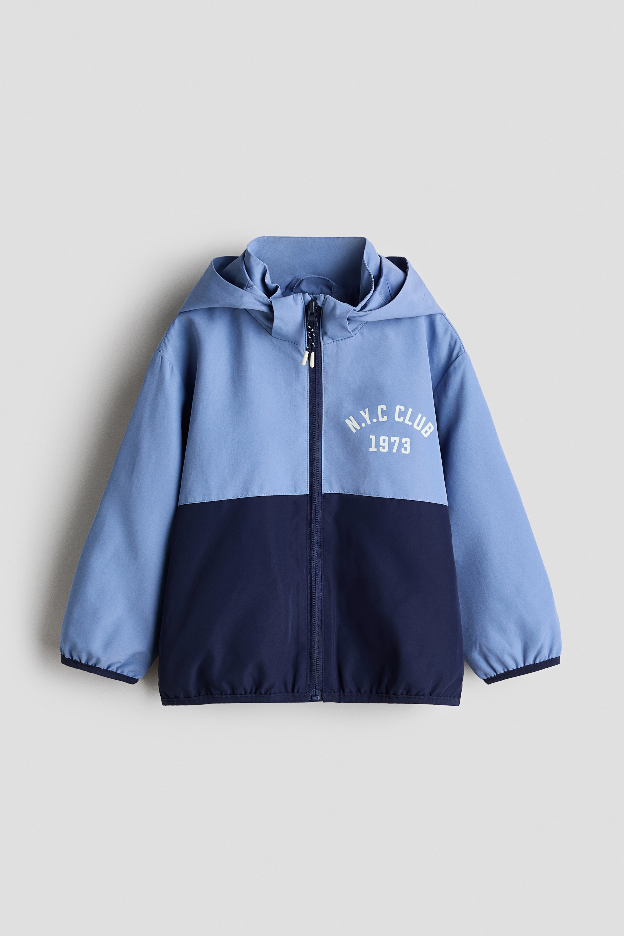 Ver imagen más grande: Una chaqueta con capucha bicolor se presenta de frente, con la mitad superior en azul claro y la mitad inferior en azul oscuro. Muestra un cierre de cremallera a toda longitud y el texto blanco "N.Y.C CLUB 1973" en el pecho izquierdo.