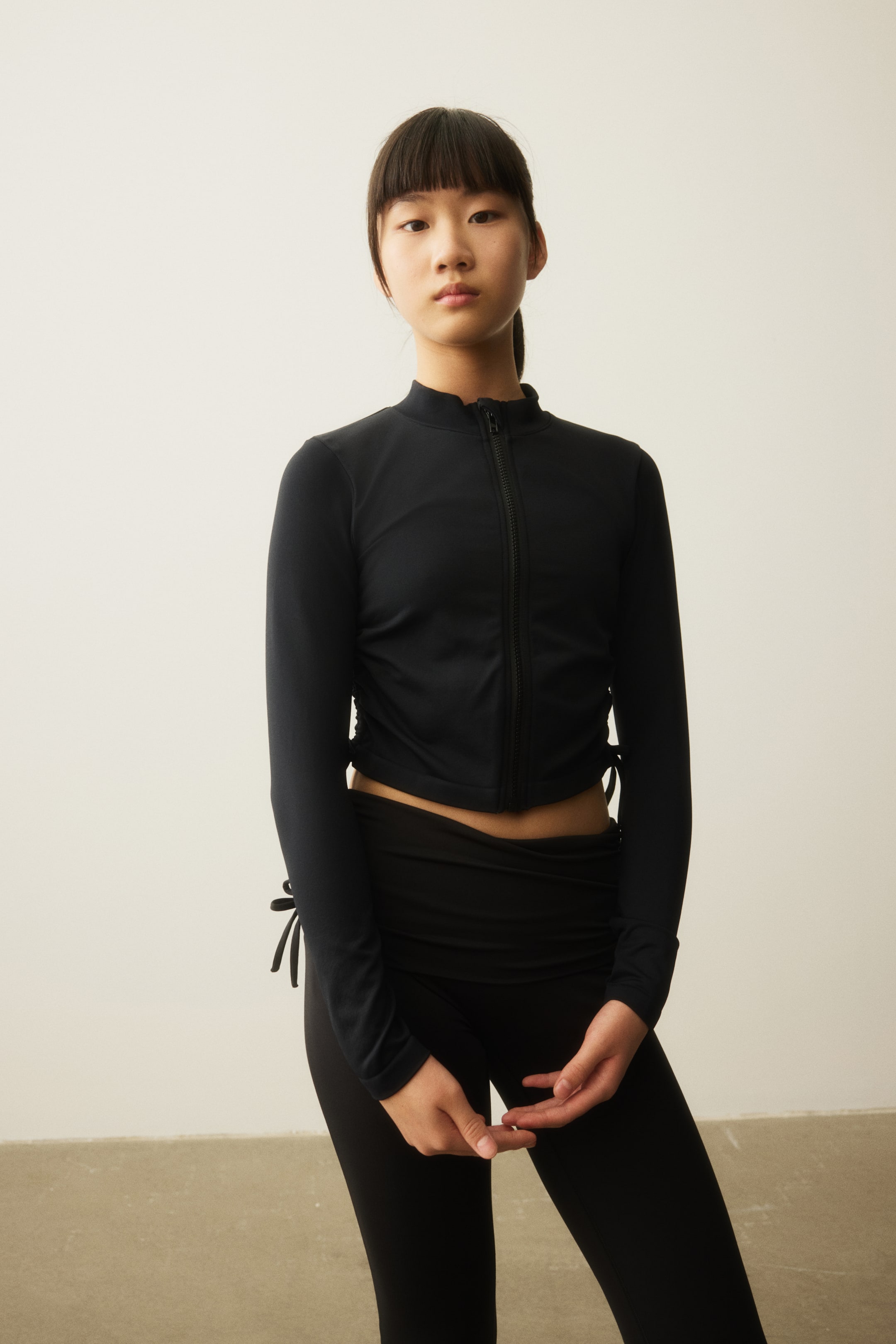 拡大画像を表示: 黒の長袖モックネックトップ（フロントにフルレングスのジッパー、サイドに調節可能なドローストリング付き）を、黒のハイウエストレギンスと合わせて着用しています。