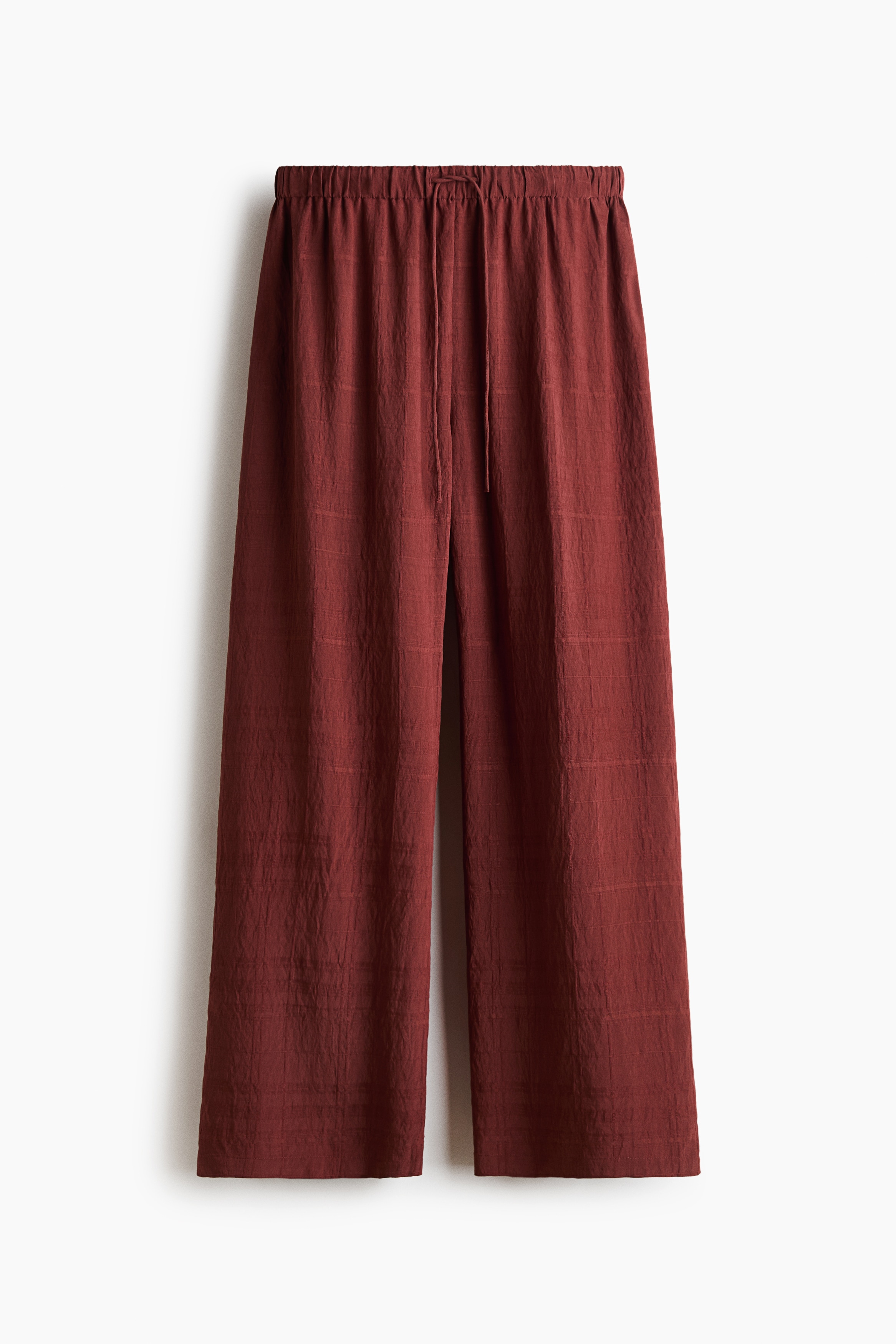 Reddish-brown wide-leg trousers are shown from the front, featuring an elasticated waistband with a thin, matching drawstring. The trousers drape straight to highlight their loose fit and textured weave fabric.