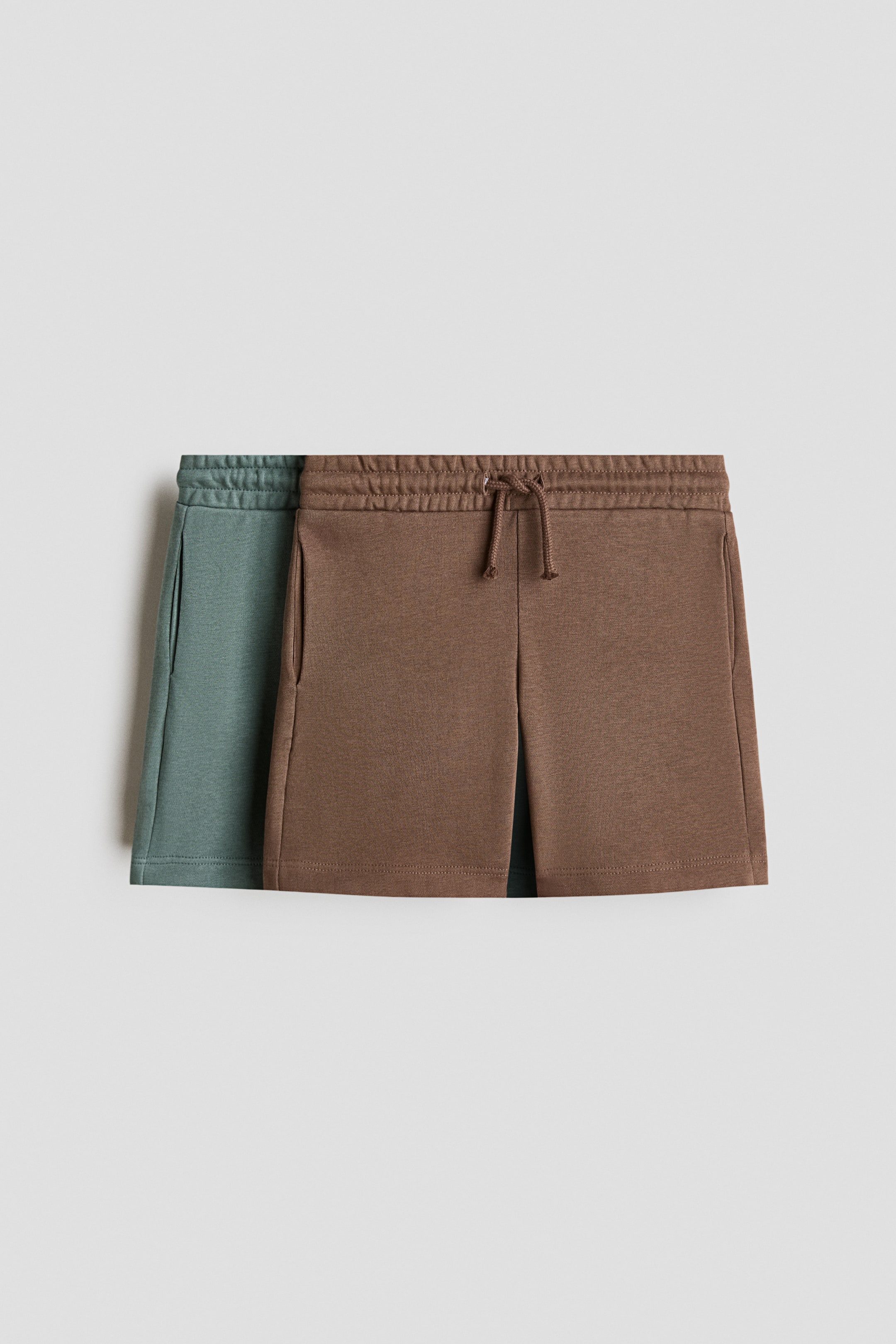 View larger image: Two pairs of shorts in lightweight sweatshirt fabric are laid out flat, viewed from a front-facing angle. The brown pair, showcasing an elasticated drawstring waist with visible drawstrings tied, and subtle side pockets, partially overlaps a teal pair with identical design features.