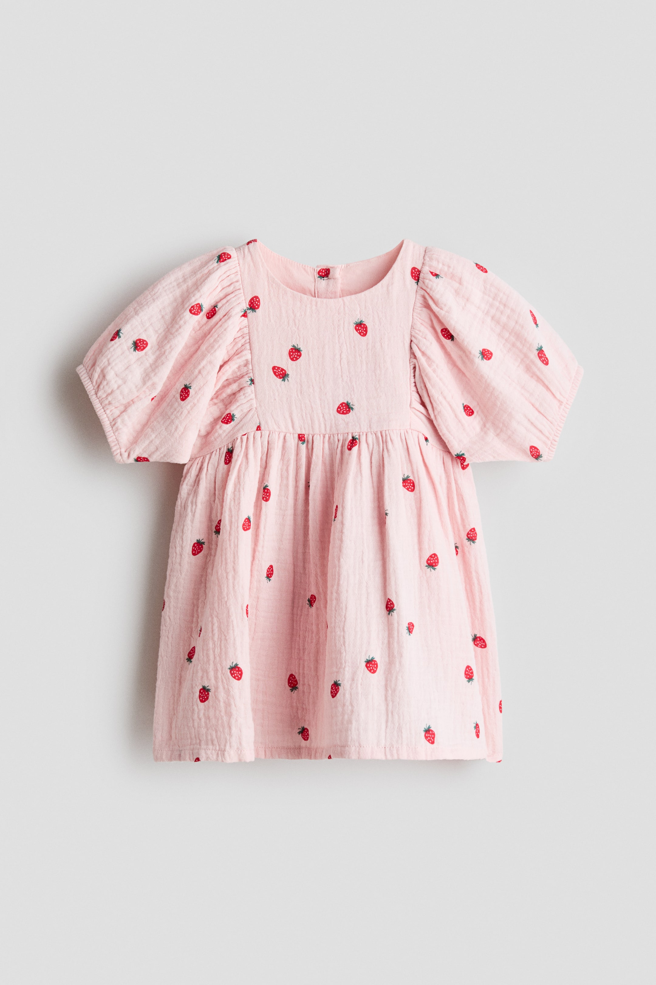 Agrandir l'image: Une robe rose clair, vue de FACE, présente un tissu en coton seersucker texturé avec un imprimé fraise dispersé. Elle est conçue avec un col rond, un empiècement étroit et des manches courtes bouffantes volumineuses, se rassemblant à la taille pour créer une jupe légèrement évasée. Des boutons sont visibles à l'arrière du col.