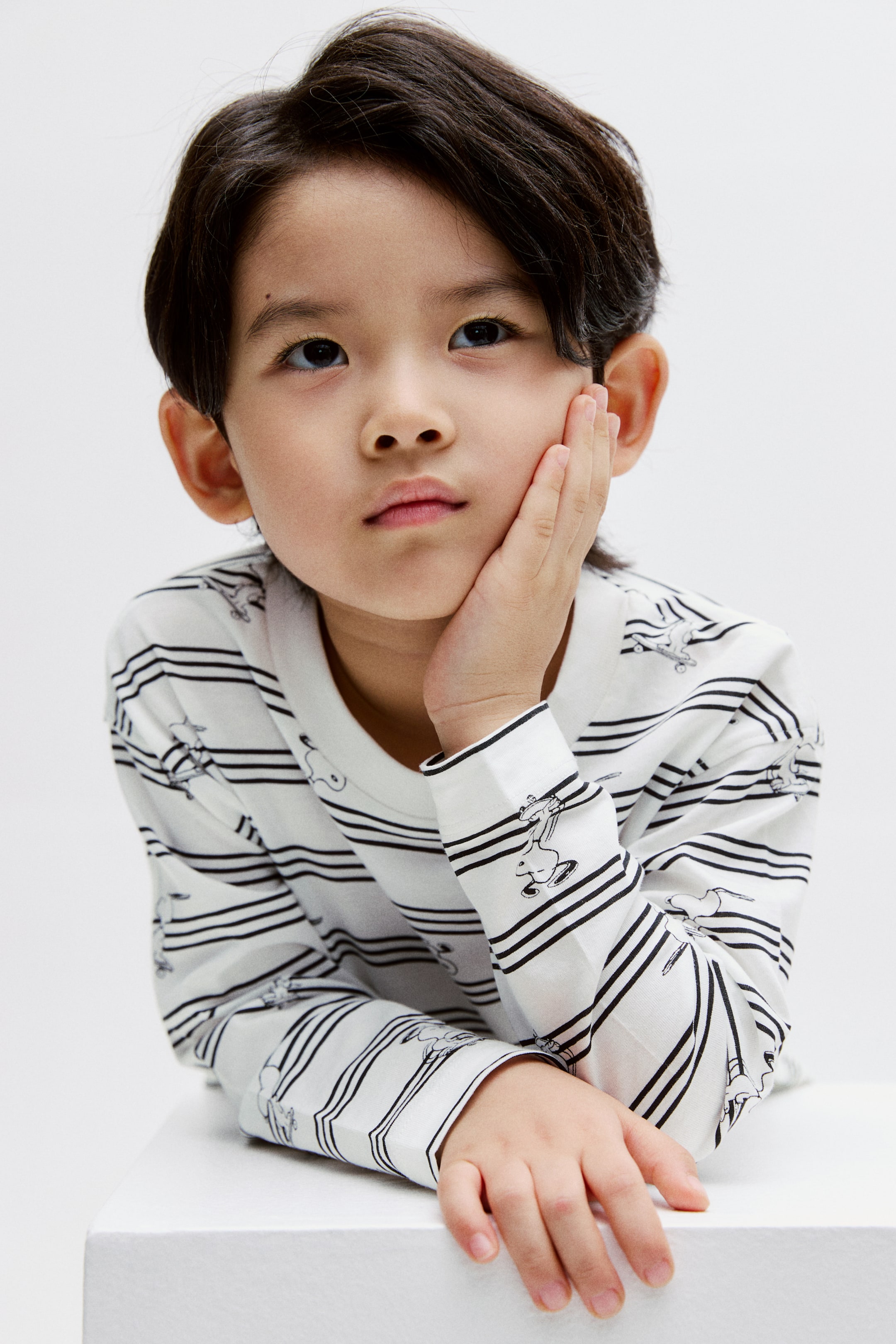 拡大画像を表示: 白い長袖シャツが着用されています。薄い黒の水平ストライプと小さな黒のスタイライズされた漫画のキャラクターが特徴です。クルーネックで、袖口が仕上げられた長袖です。