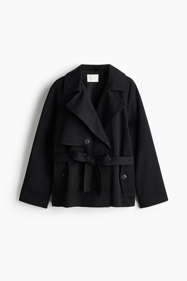 A black short trench coat showcases a double-breasted front with wide notch lapels. It features a matching fabric belt tied in a bow at the waist, two buttoned flap pockets, and storm flaps across the shoulders. - 1
