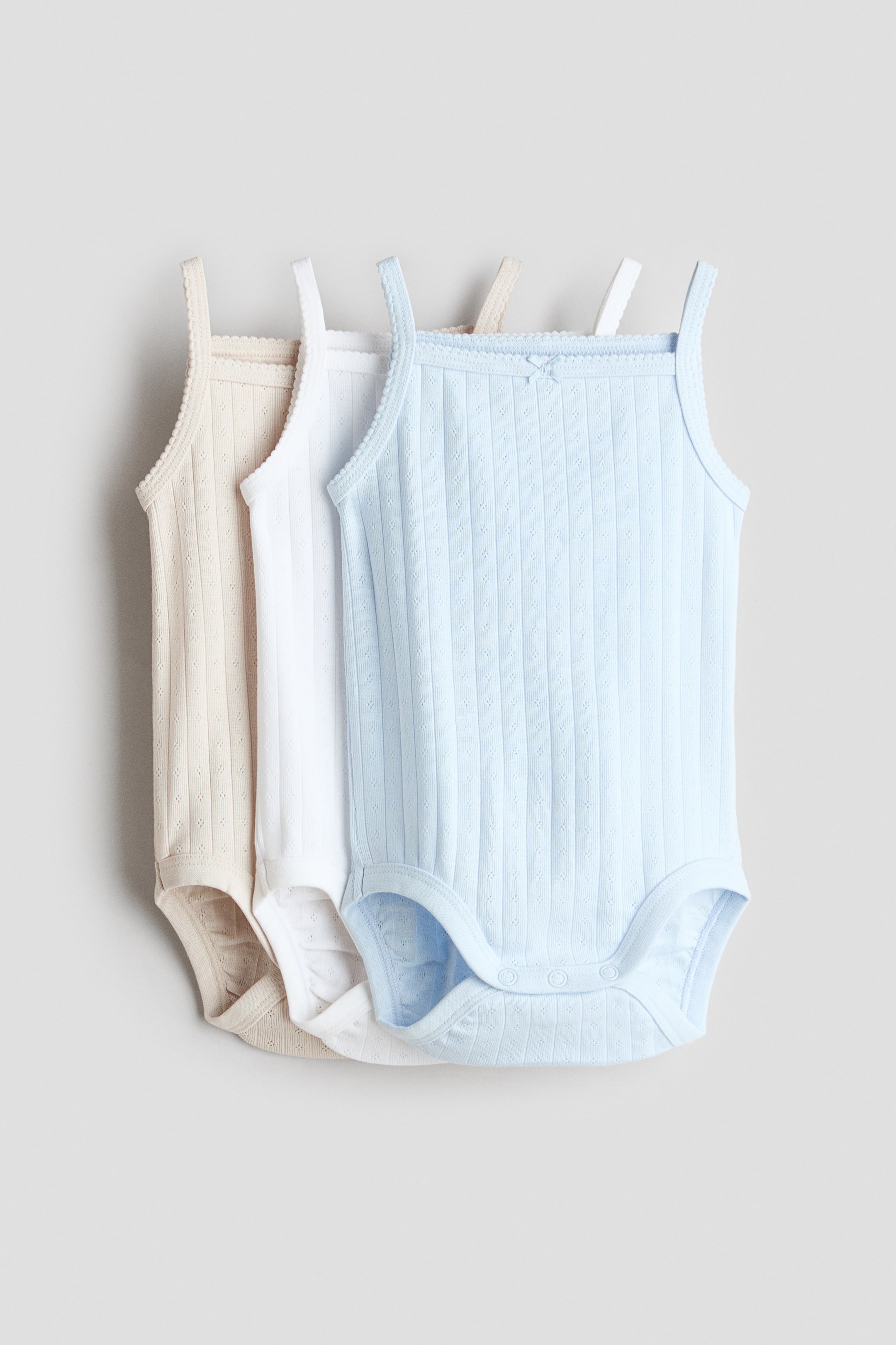 View larger image: Sleeveless bodysuits are showcased from the front in a set of three, available in light blue, white, and light beige. Each bodysuit features a ribbed cotton jersey with eyelet patterns, slender shoulder straps, a square neckline with scalloped trim and a small decorative bow, and press-studs at the crotch.