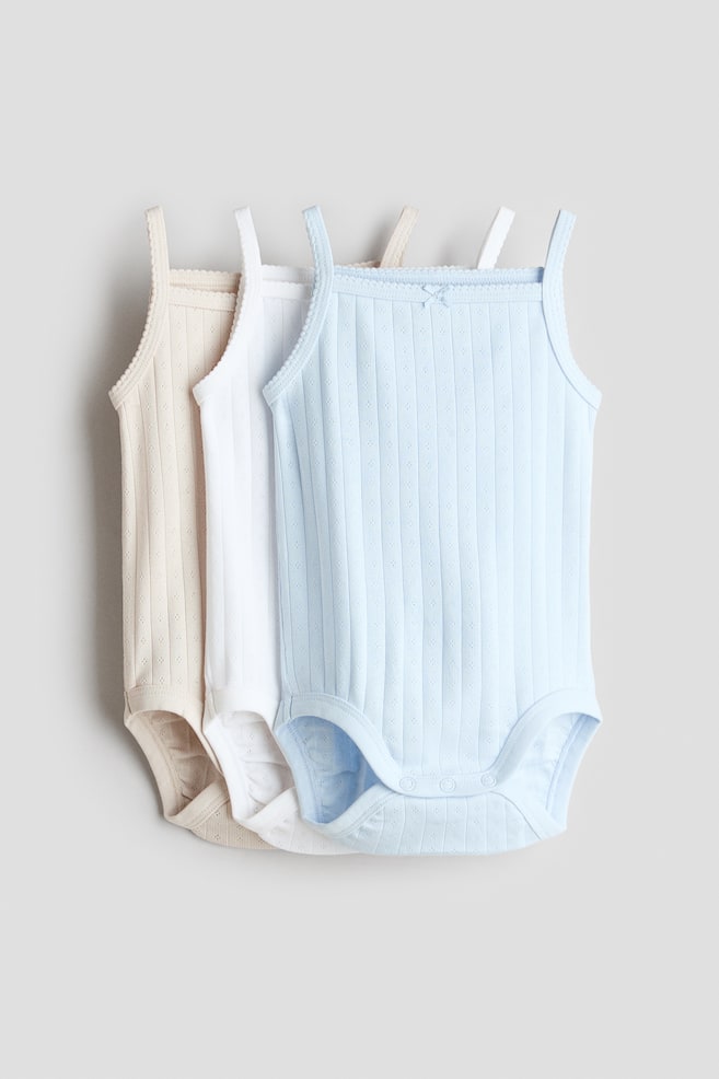 Sleeveless bodysuits are showcased from the front in a set of three, appearing in light blue, white, and light beige. Each bodysuit features a ribbed cotton jersey with eyelet patterns, slender shoulder straps, a square neckline with scalloped trim and a small decorative bow, and press-studs at the crotch. - 1