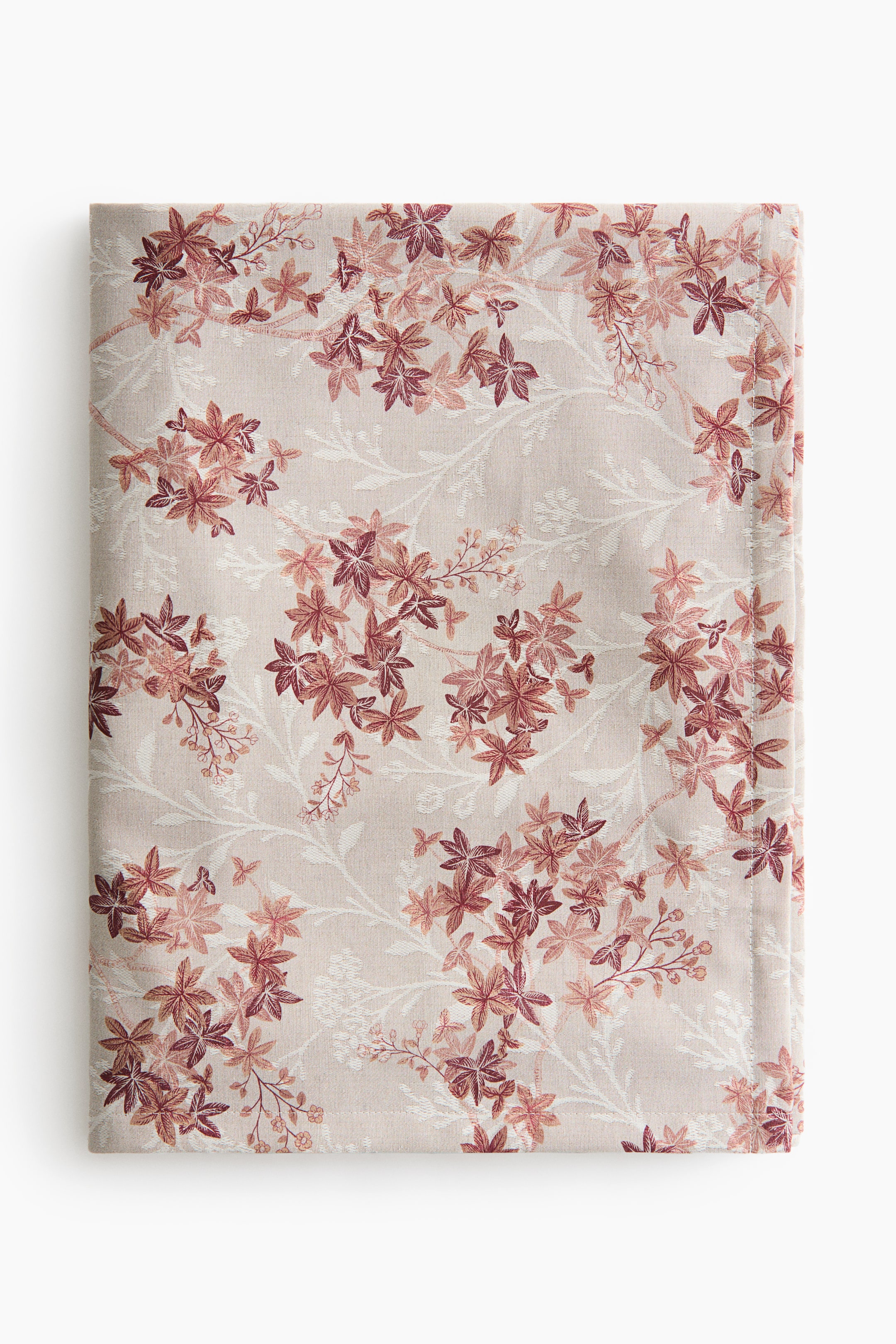 View larger image: A folded tablecloth displays a repeating botanical pattern of reddish-brown and muted pink leaf clusters on delicate branches over a light beige background. The textile is presented in a flat front view, showcasing the intricate details of the warm-toned leaves and subtle white floral motifs woven throughout.