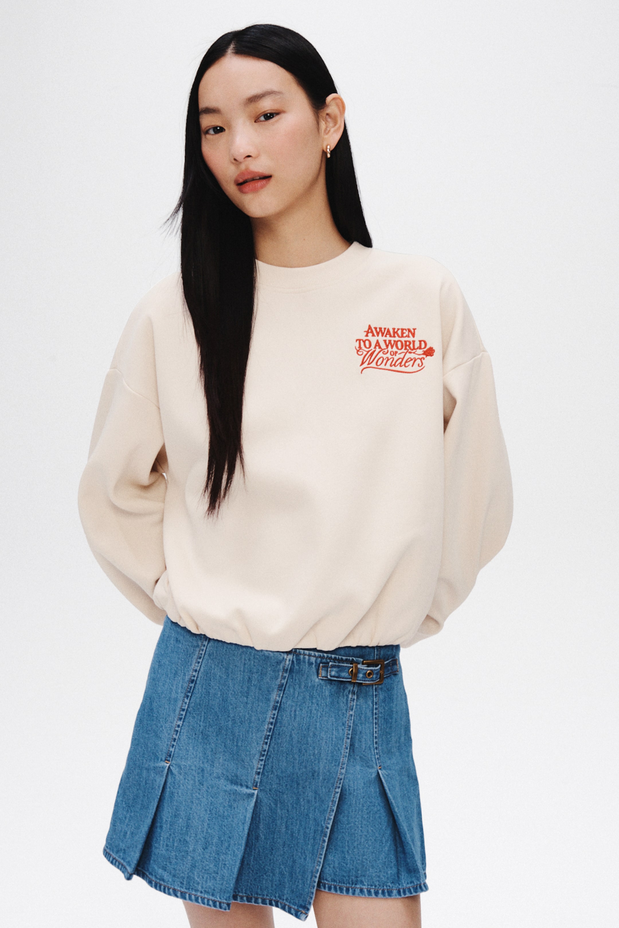 View larger image: A cream-colored sweatshirt is shown from the front, featuring a crew neck, long sleeves, and a gathered hem that creates a bloused silhouette. Red embroidered text reading "Awaken to a world of wonders," accompanied by a small flower graphic, appears on the left chest.