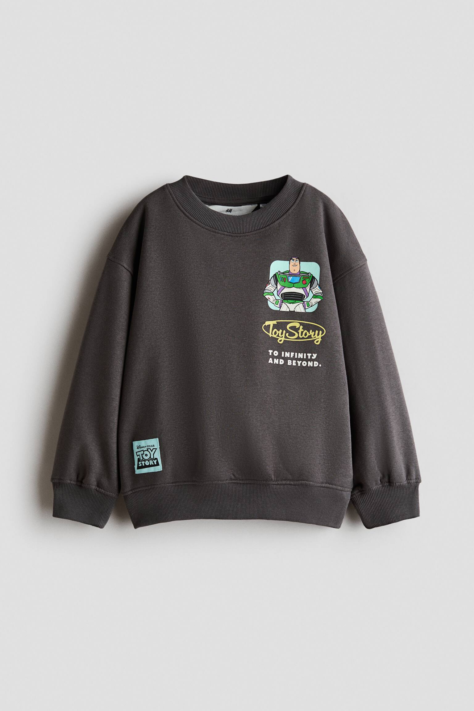 A dark grey sweatshirt is shown from the front, featuring a rib-trimmed crew neckline, dropped shoulders, and long sleeves with ribbed cuffs. The front displays a printed graphic of Buzz Lightyear with 'Toy Story' text on the chest, along with a small light blue patch near the ribbed hem.