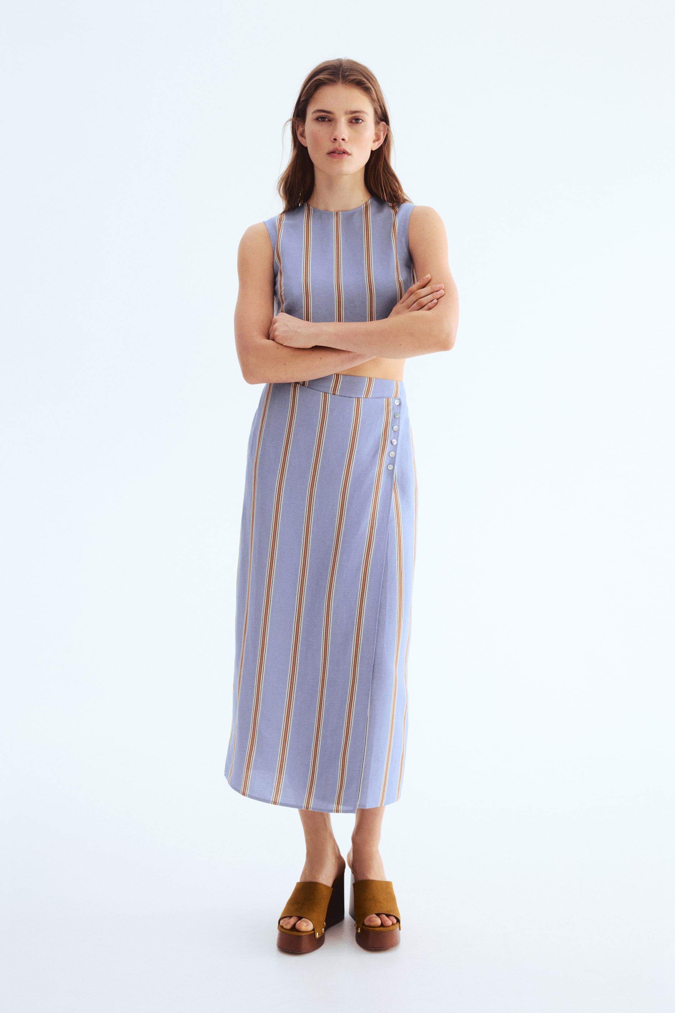 View larger image: A coordinated two-piece ensemble showcasing a sleeveless, crew-neck crop top paired with a midi skirt. The blue ensemble is adorned with vertical stripes in white and brown, with the skirt featuring an overlapping front panel fastened by light-coloured buttons on the right side.