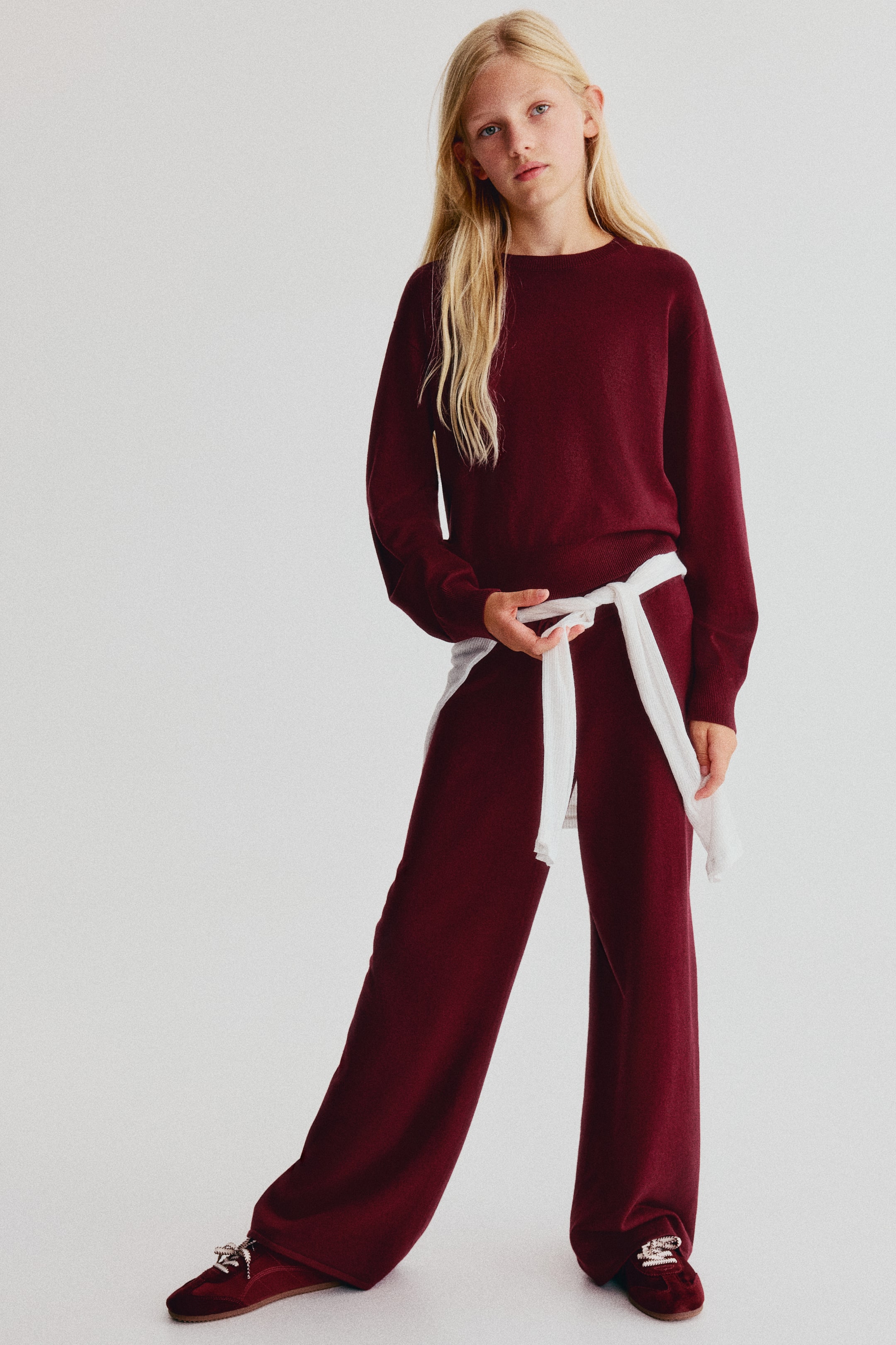 Grösseres Bild ansehen: Ein Set aus dunkelrotem Feinstrickpullover und Jogginghose ist von vorne zu sehen. Es besteht aus einem Langarm-Pullover mit rundem, geripptem Ausschnitt, überschnittenen Schultern und gerippten Bündchen, kombiniert mit einer passenden Jogginghose mit geradem Bein. Ein kontrastierender weisser Strickpullover ist um die Taille der Jogginghose gebunden und rundet den gestylten Look ab.