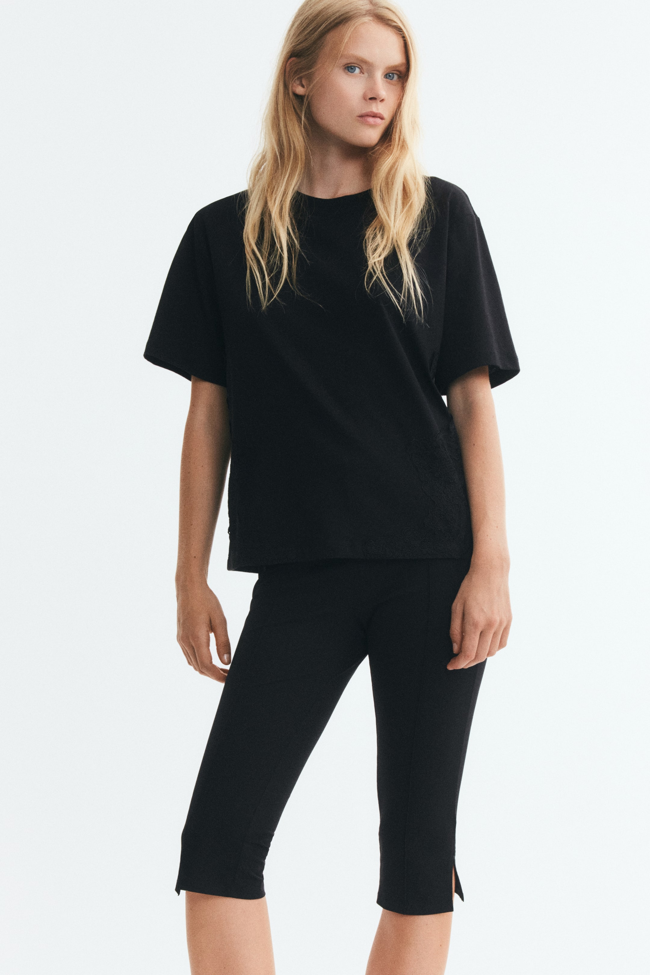 View larger image: A black, loose-fit T-shirt featuring a round neckline, dropped shoulders, and subtle lace inserts along the lower sides is being worn by the model. The T-shirt is styled with matching black slim-fit cropped trousers.
